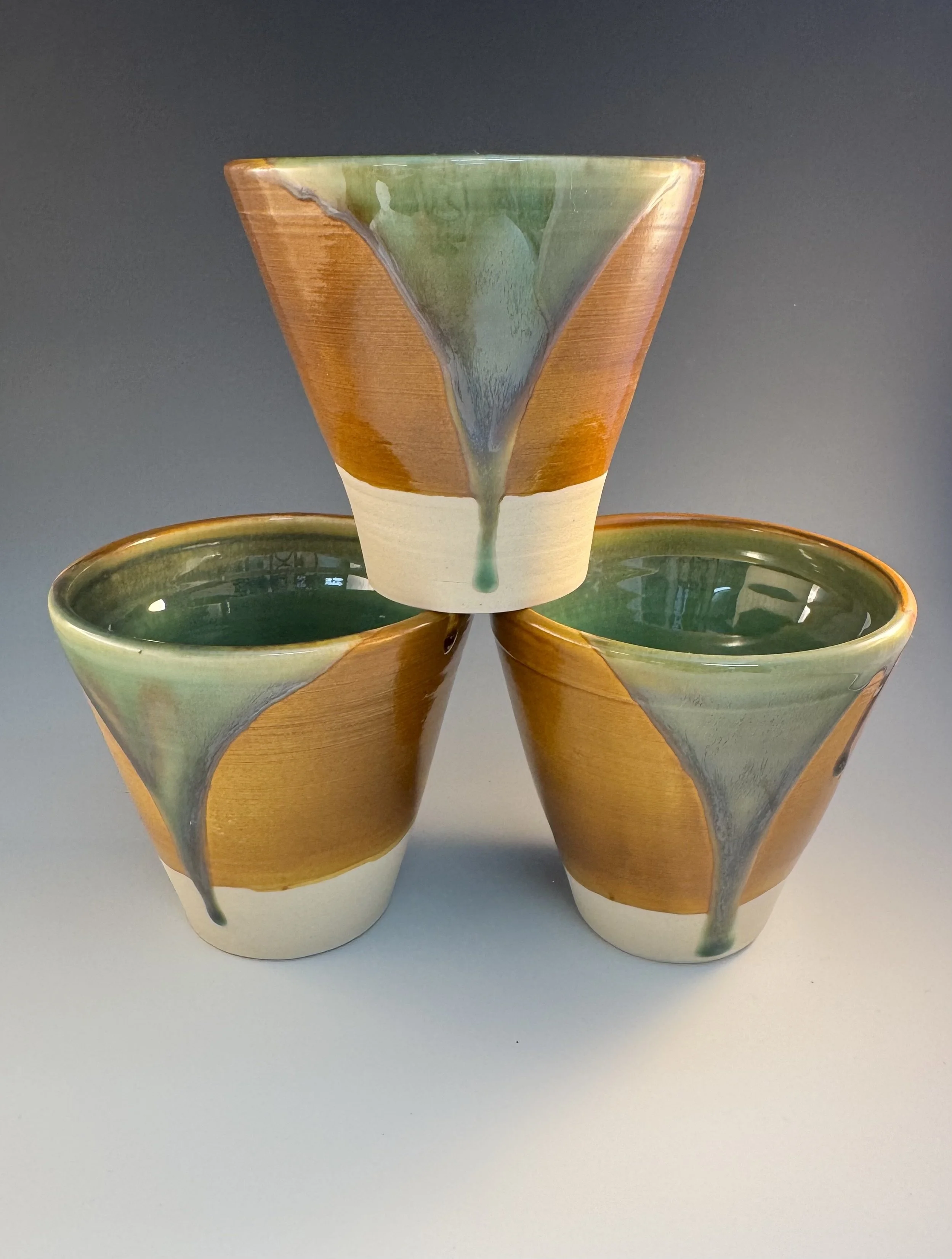 Three ceramic cups with a glaze that features green, brown, and cream colors, stacked in a pyramid formation on a grey surface with a dark background.