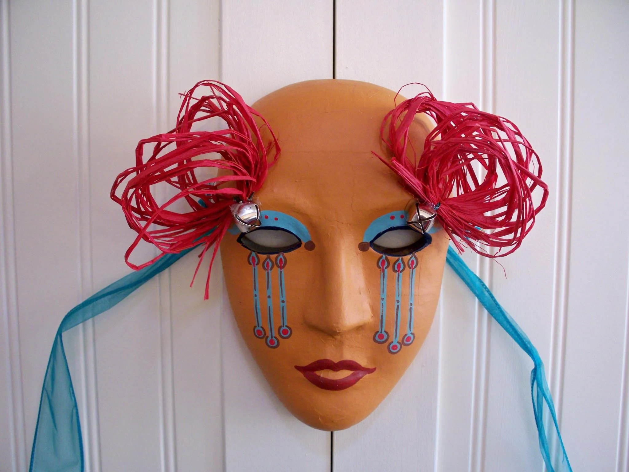A decorative mask with a peach-colored face, blue eyeshadow, red lips, and long eyelashes. Red and blue ribbon accents are attached to the mask, which is mounted on a vertical white panel wall.
