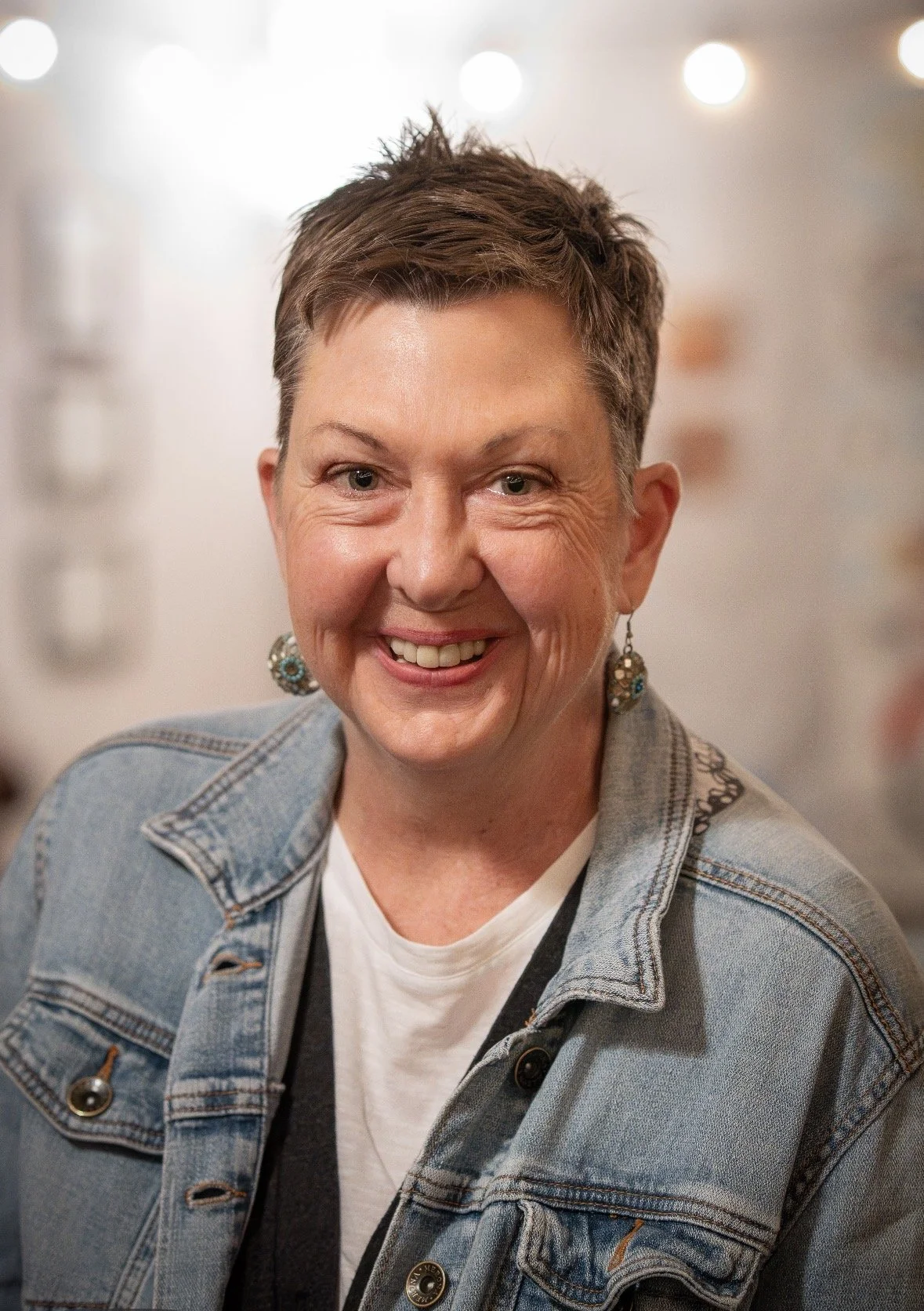 Close-up of a smiling woman with short brown hair, wearing a denim jacket, white t-shirt, and turquoise earrings, with lights in the background.
