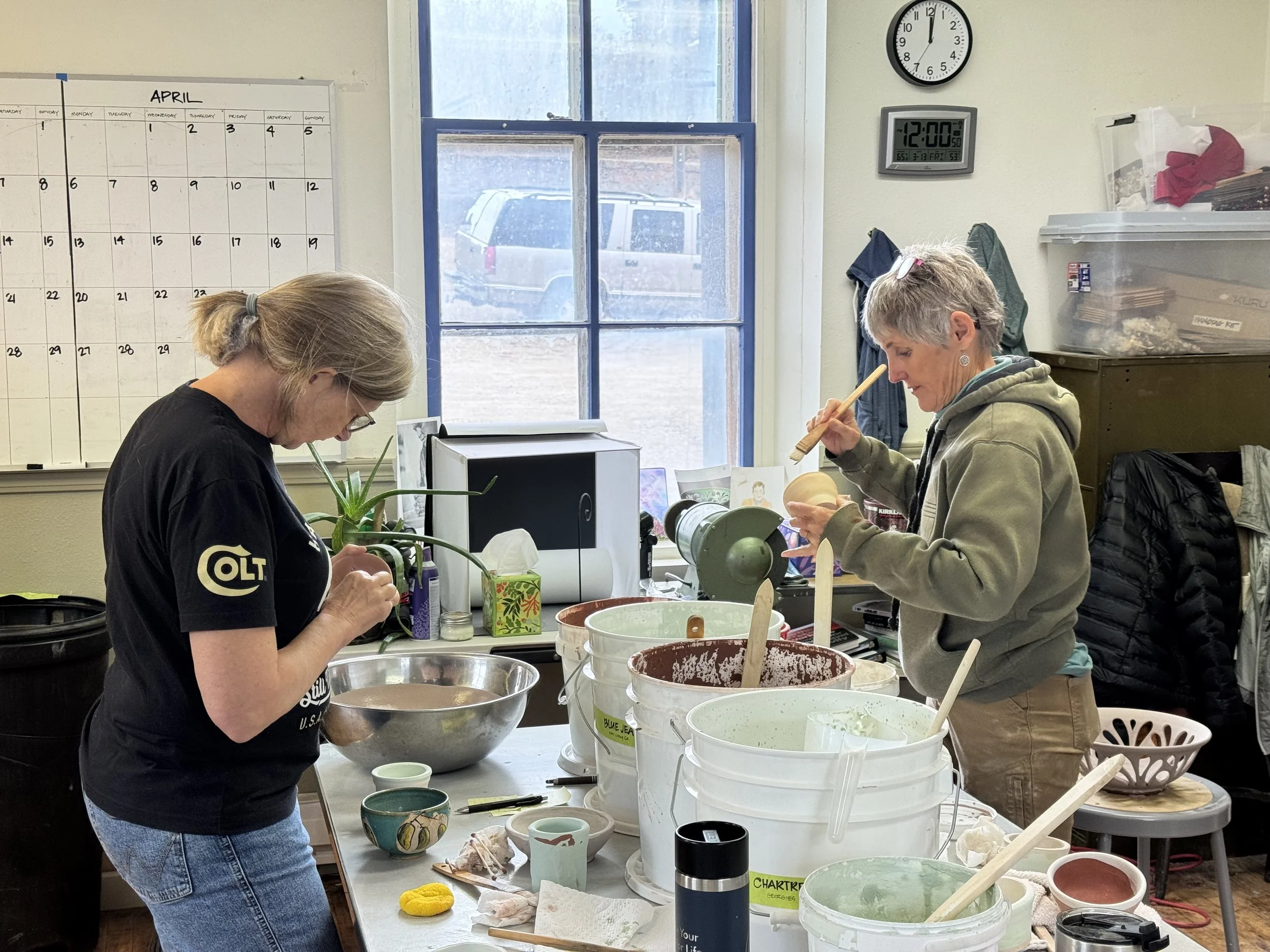 Two women working on pottery projects at a table filled with bowls and clay in a ceramics studio. One woman is using a tool to shape clay, while the other is holding a piece of pottery and working on it.