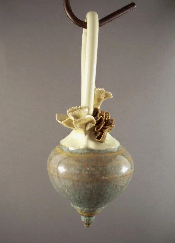 A ceramic pendant with a small hanging container, holding mixed wood shavings, with a loop at the top for hanging.