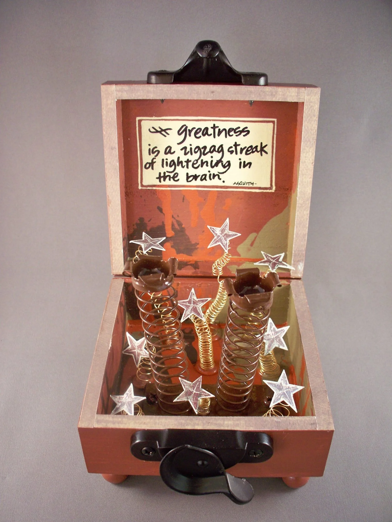 A wooden box with a quote on the inside back wall that reads, 'Greatness is a zigzag streak of lightening in the brain.' The box contains decorative items including star-shaped ornaments, coil springs, and small brown objects, all positioned on a gray surface.