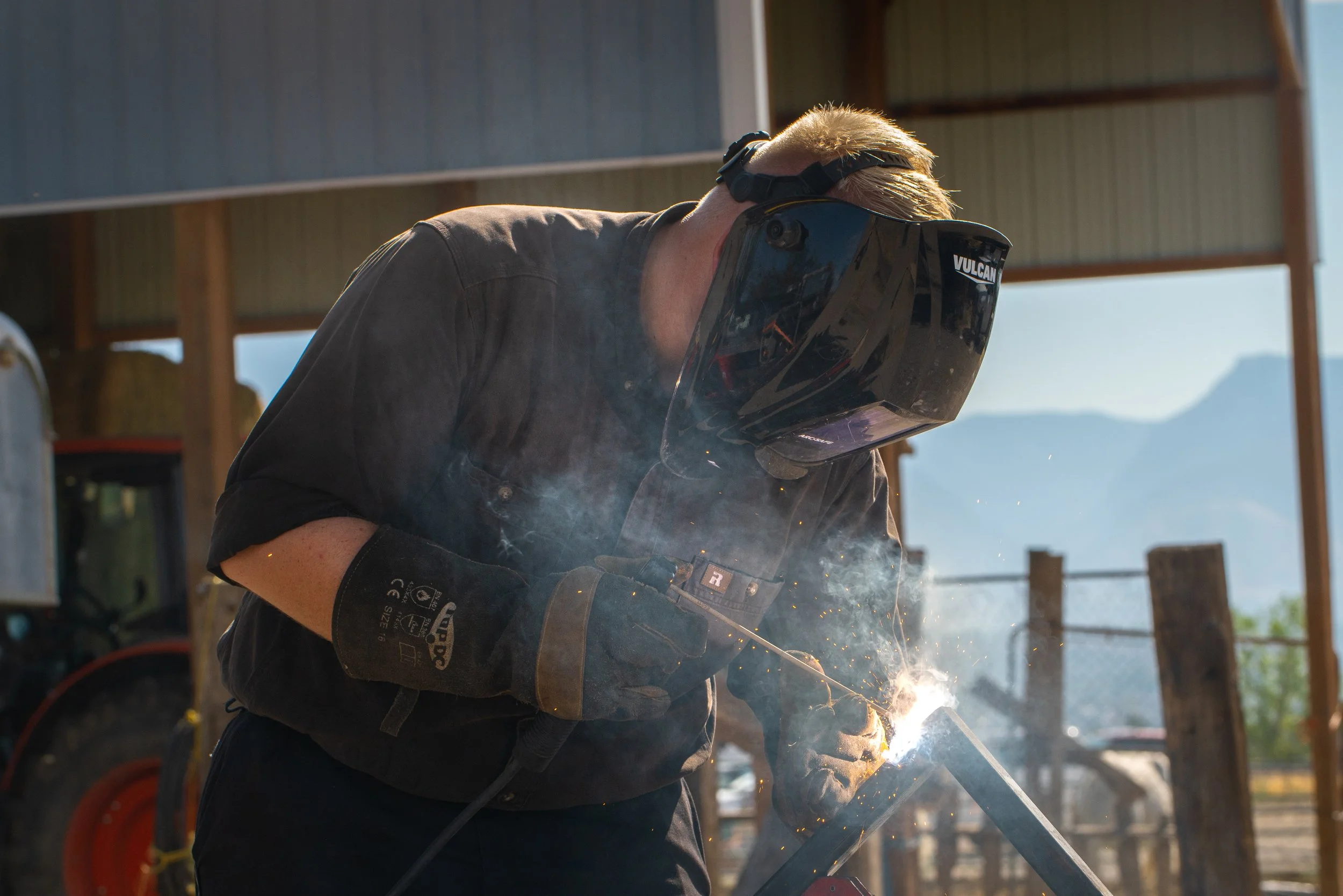 Person welding outdoors, wearing protective gear including a welding helmet and gloves, sparks flying as they work on metal.