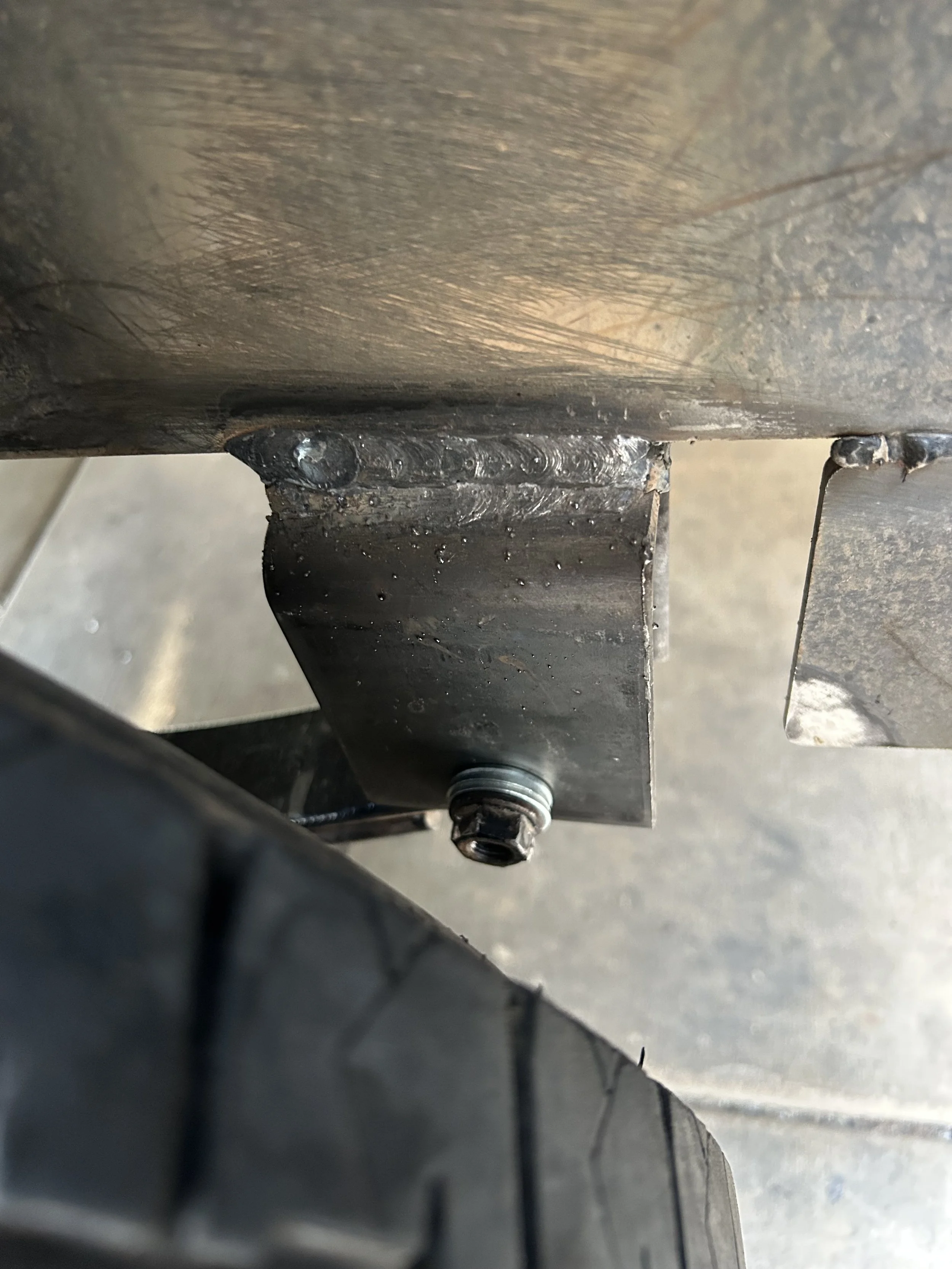 Close-up of a medium-sized metallic object with a welded connection and a bolt at the bottom, showing a surface with scratches and slight rust, possibly part of a mechanical or industrial device.