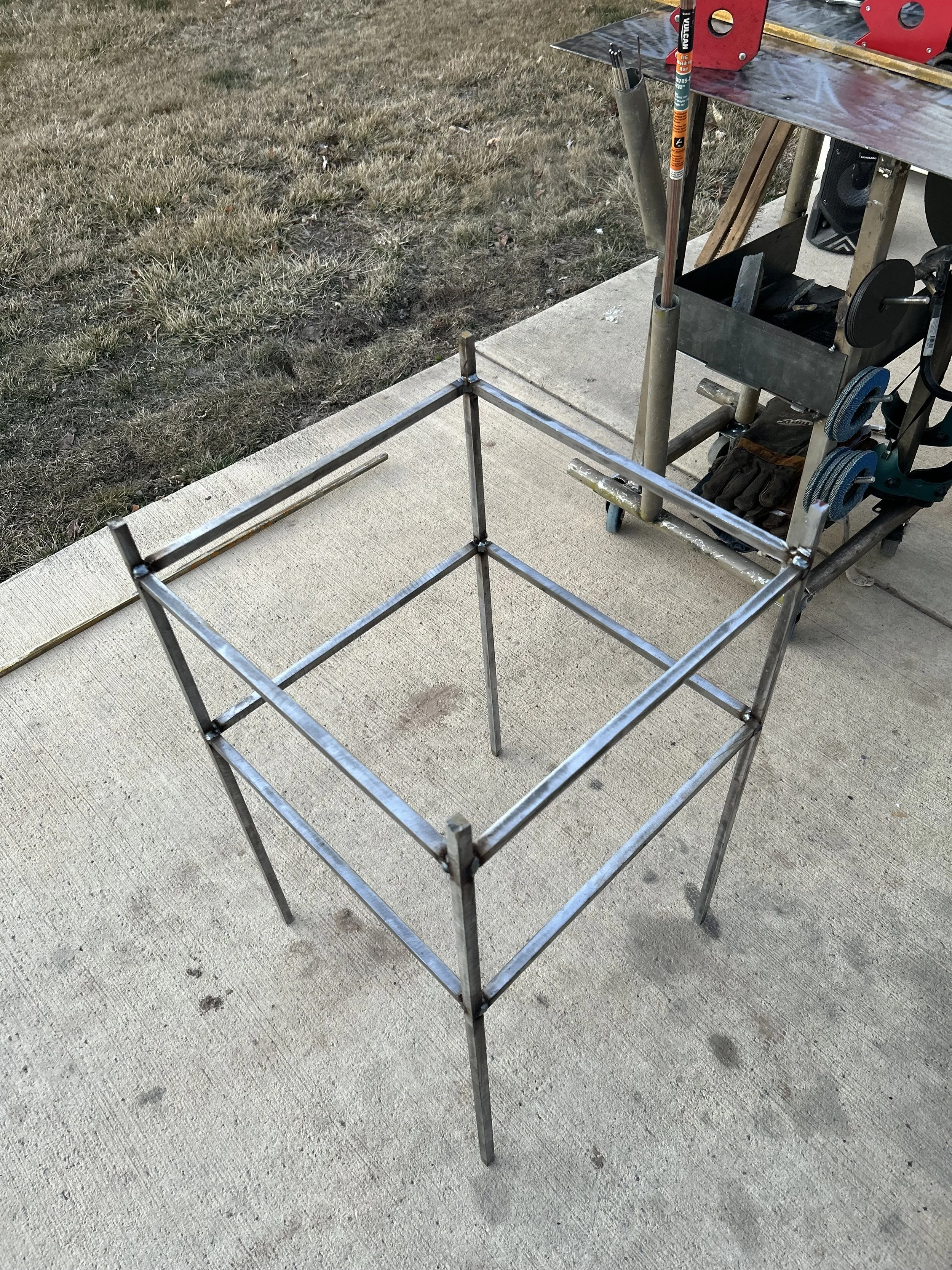 Metal framework structure placed on a concrete sidewalk with grass in the background, part of a worksite or DIY project.