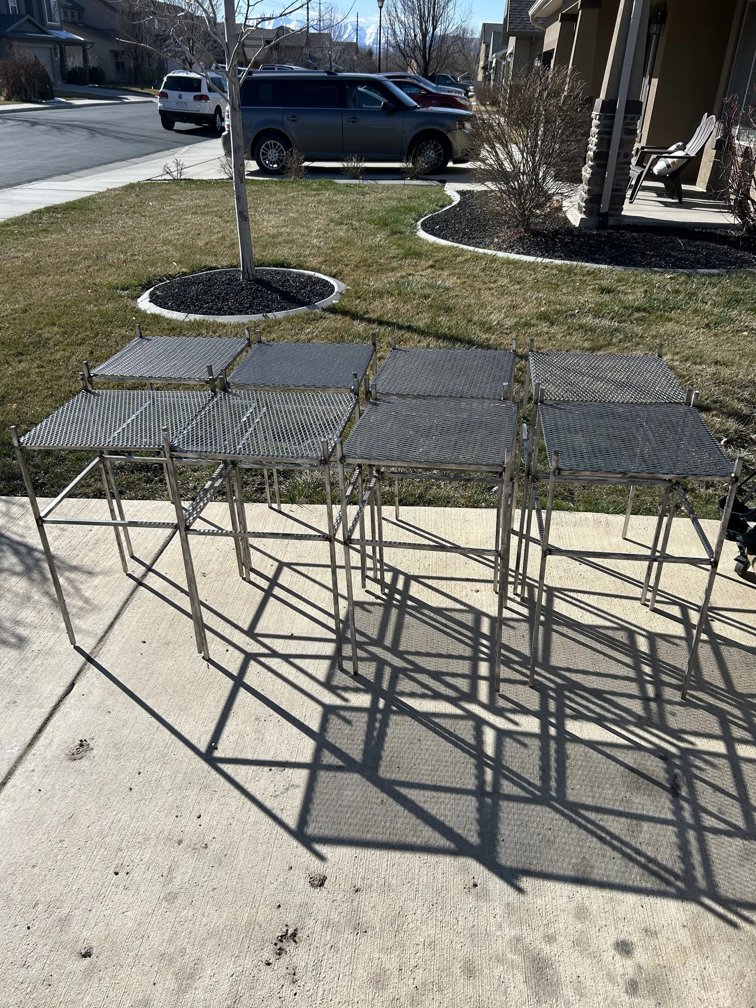 Metal outdoor furniture on a concrete patio with shadow reflecting on the ground, small paw prints on the concrete, a yard with grass, a tree in a circular bed, and cars parked on the street in a residential neighborhood.