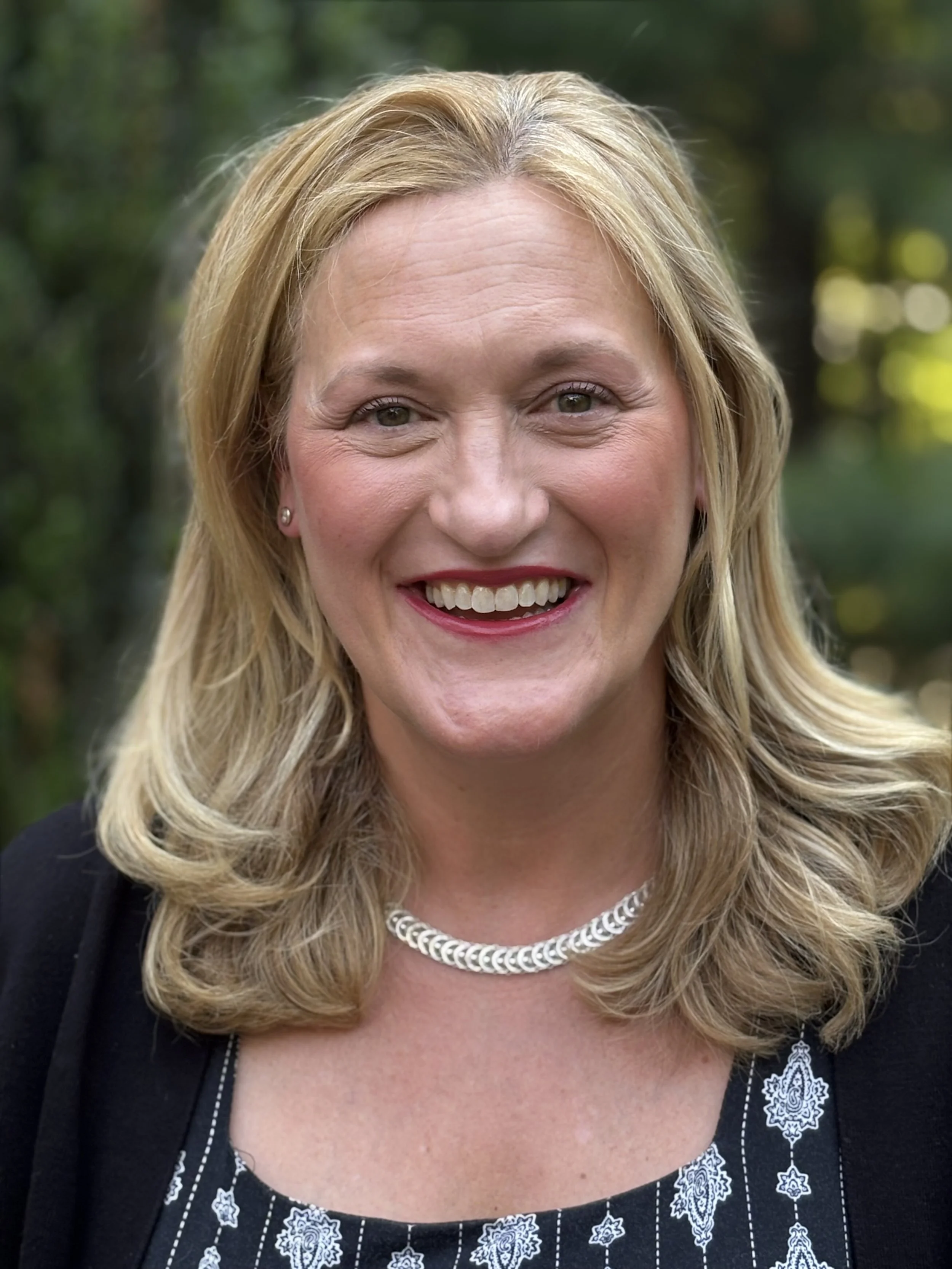 A woman with blonde hair smiling outdoors, wearing a black jacket, a silver necklace, and pearl earrings, with a background of green blurred trees.