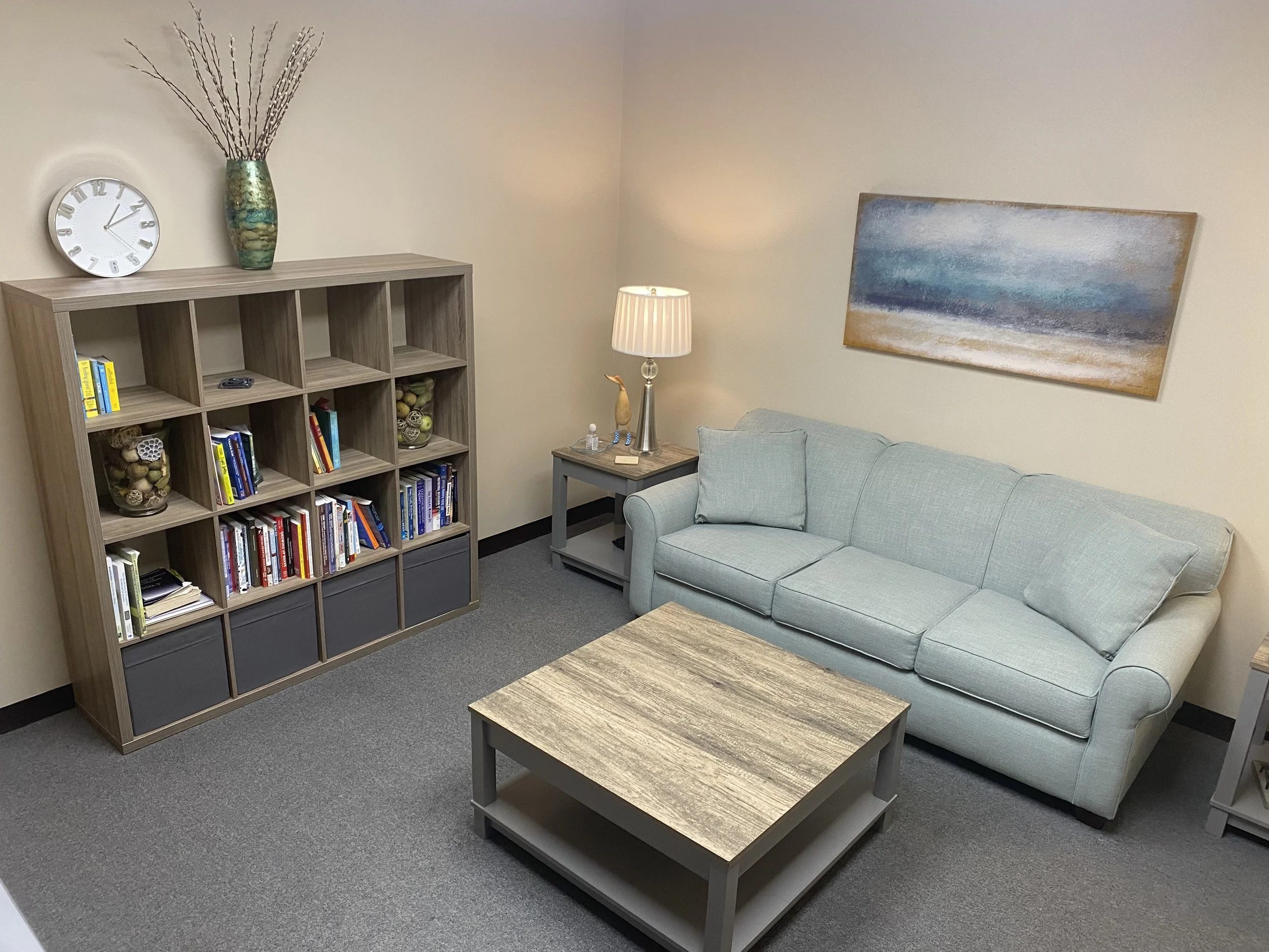 A cozy living room with a light blue sofa, a wooden coffee table, a bookshelf filled with books and decorative items, a table lamp with a beige shade, a large abstract painting on the wall, and a decorative vase with branches on top of the bookshelf.