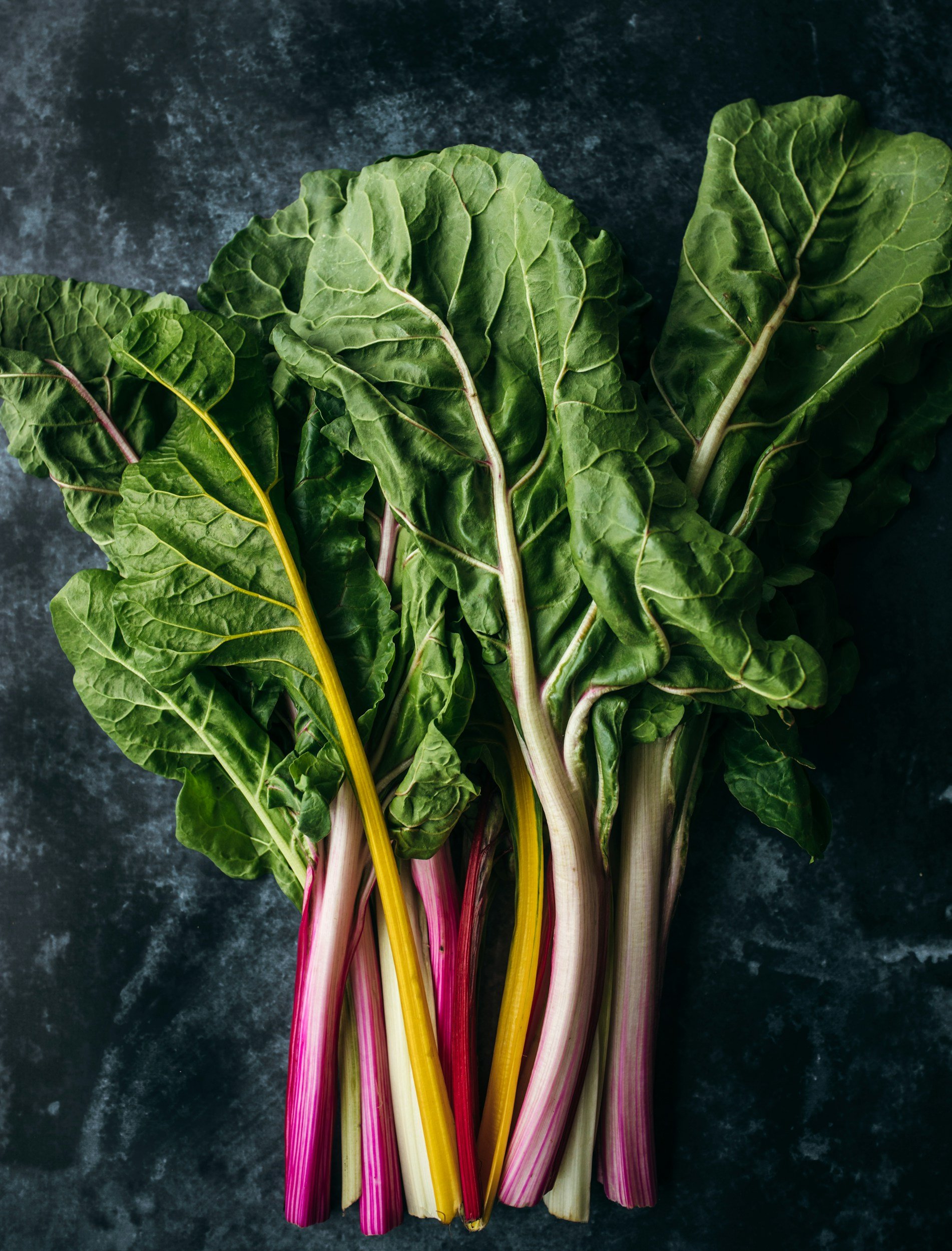 Colorful chard leaves with pink, yellow, and white stems on a dark surface.