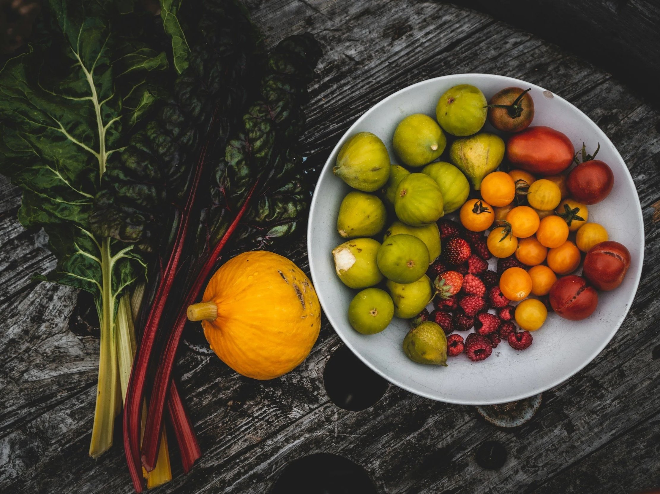 A white bowl filled with various colorful tomatoes on a weathered wooden surface, alongside dark leafy greens and a yellow pumpkin.