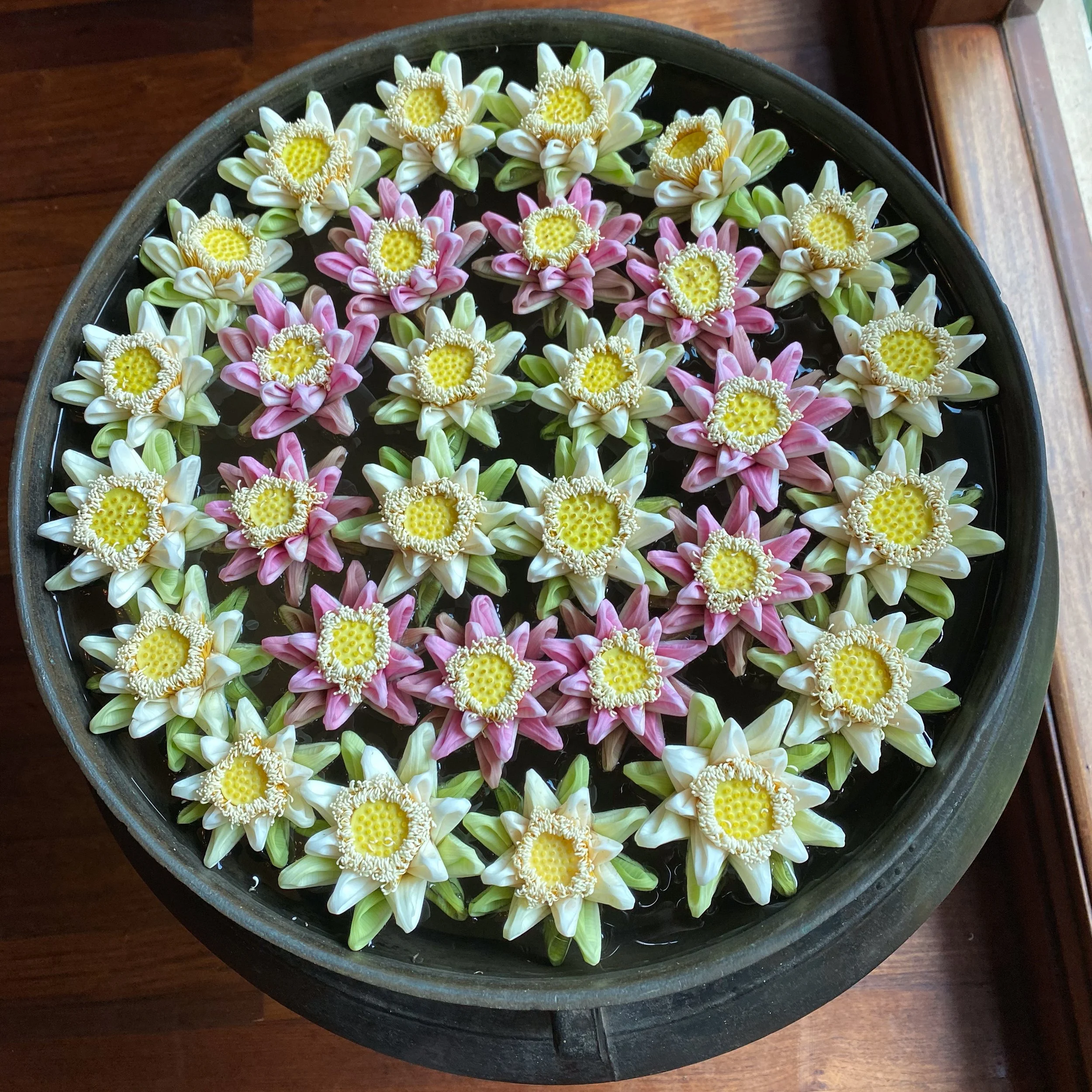 Assorted traditional flower garlands floating in a round black container filled with water, placed on a wooden surface.