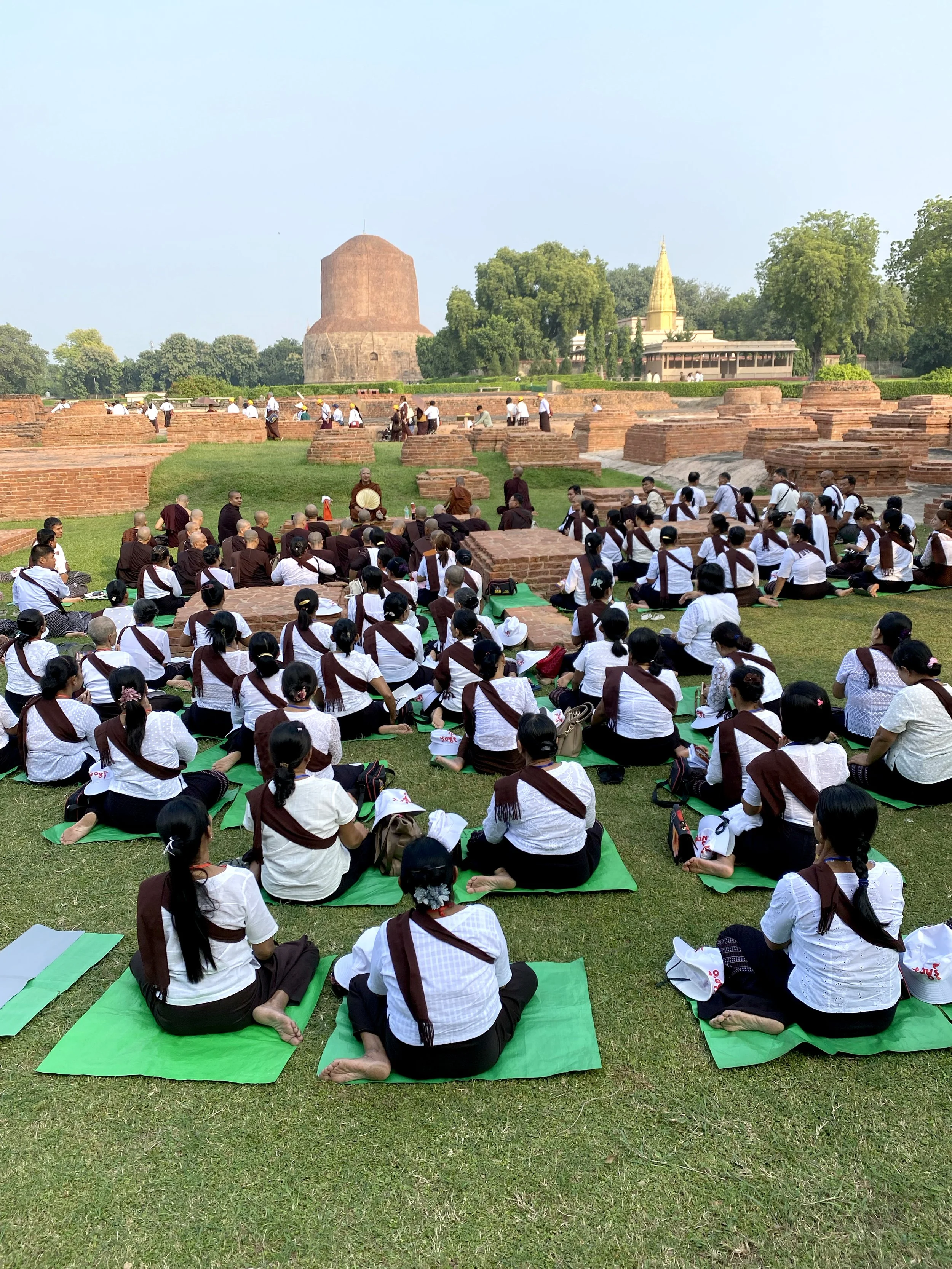 A large group of monks and laypeople sitting on green mats on a grassy courtyard, participating in a meditation or prayer session, with a historic temple and ancient brick ruins in the background.