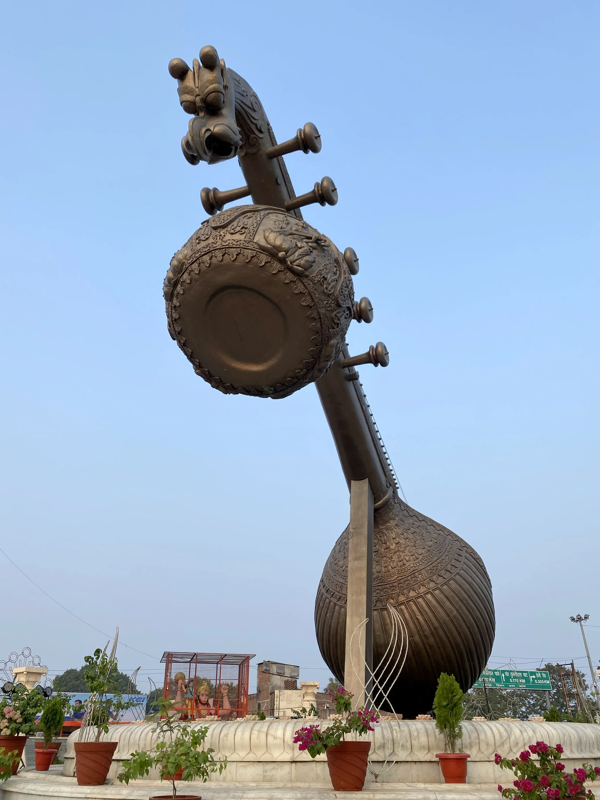 A large bronze statue of a cow with detailed carvings and musical note designs, with a bright blue sky in the background, surrounded by potted plants and flowers, and a small temple or shrine with religious figures in the foreground.