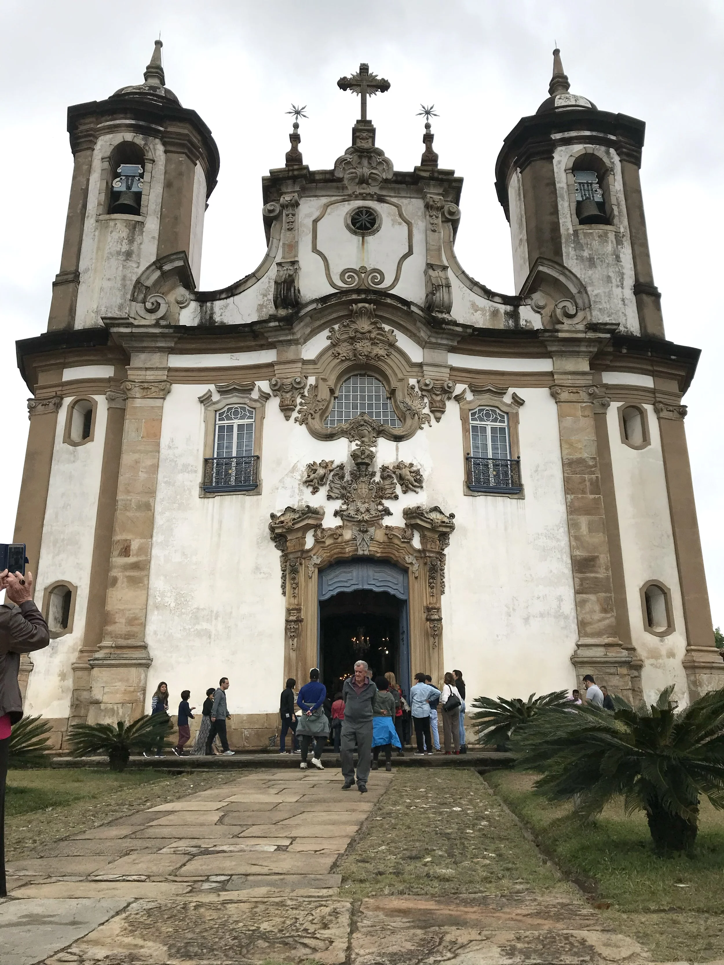 The entrance of a historic church with Baroque architecture, two bell towers, detailed sculptures, and a crowd of people outside, some taking photos.