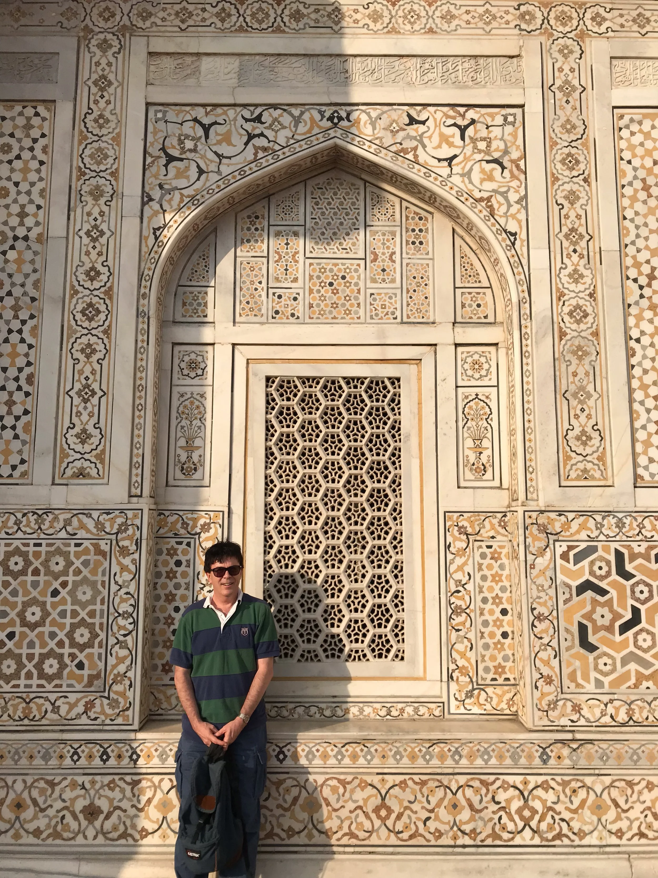A man wearing sunglasses, a green and navy striped shirt, and dark pants stands in front of an ornate, patterned wall with intricate geometric and floral designs, and a carved marble lattice window.