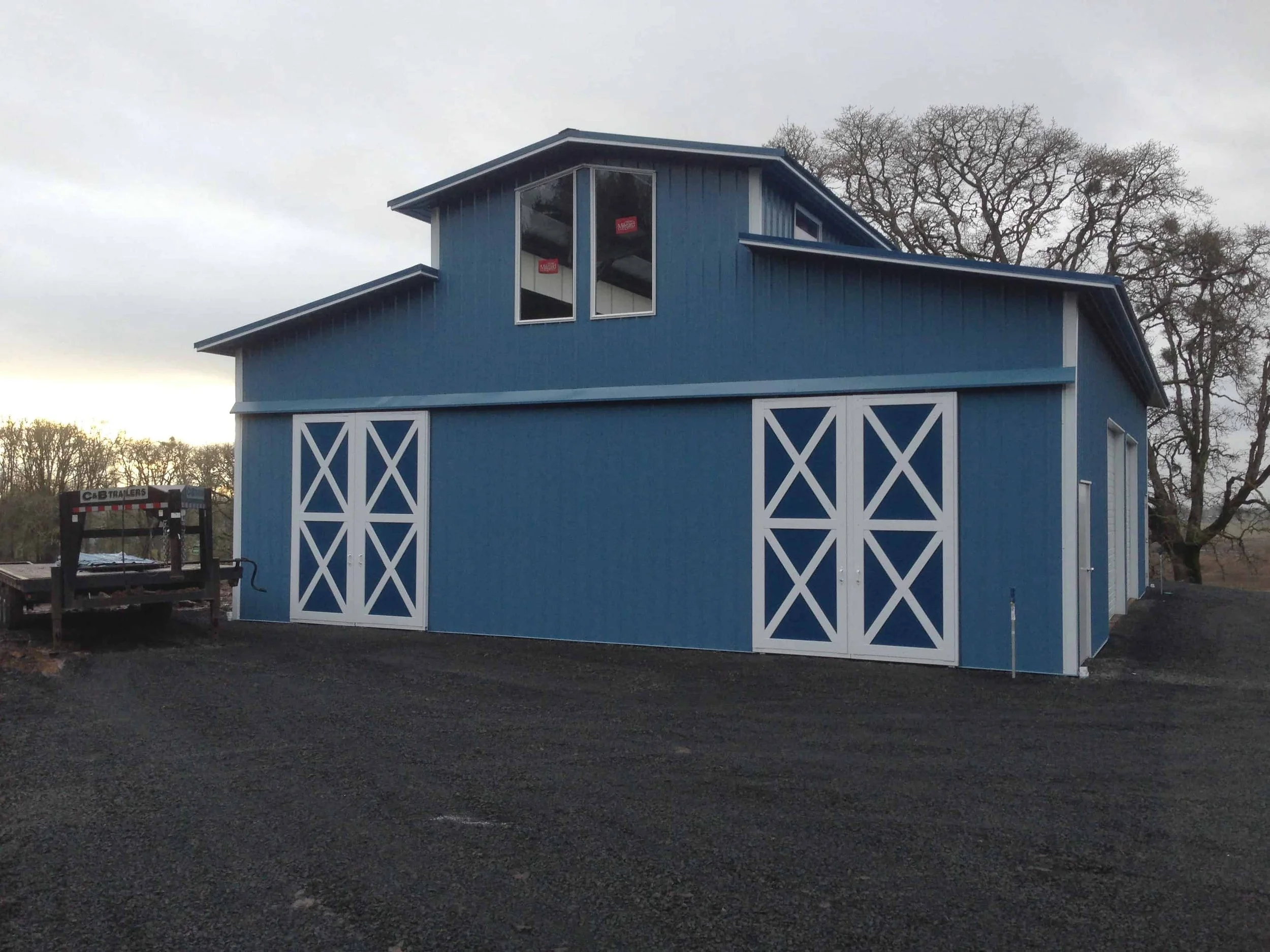 Blue barn with white double doors and white trim, set against a backdrop of bare trees on an overcast day.