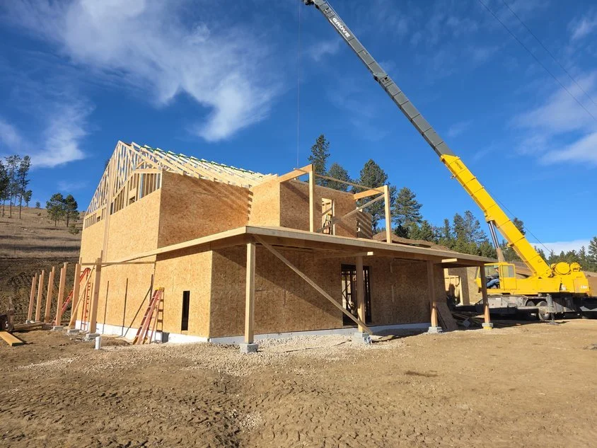 A house under construction with wooden framing and plywood sheathing, a crane lifting materials, and a clear blue sky.