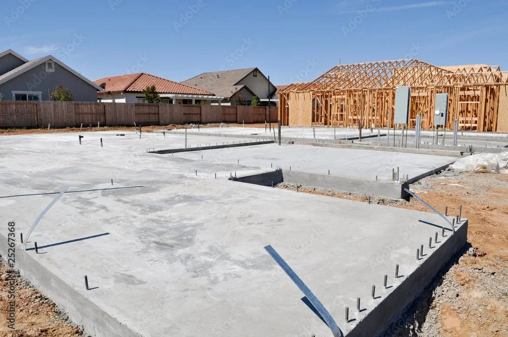 Foundation of a house under construction with concrete slab and plumbing pipes, wooden framing of a neighboring house being built, in a suburban neighborhood under a clear blue sky.