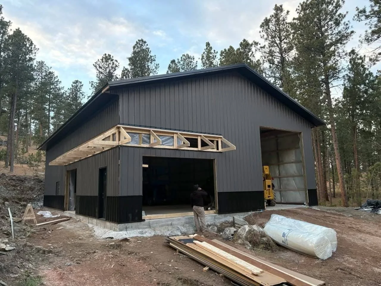 Under construction black metal building in a wooded area with construction materials and a worker in front.
