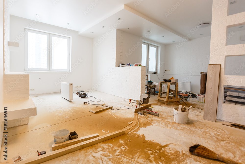 Interior of a room under renovation with construction tools and materials scattered on a plywood-covered floor, white walls, large windows, and two small windows on a side wall.