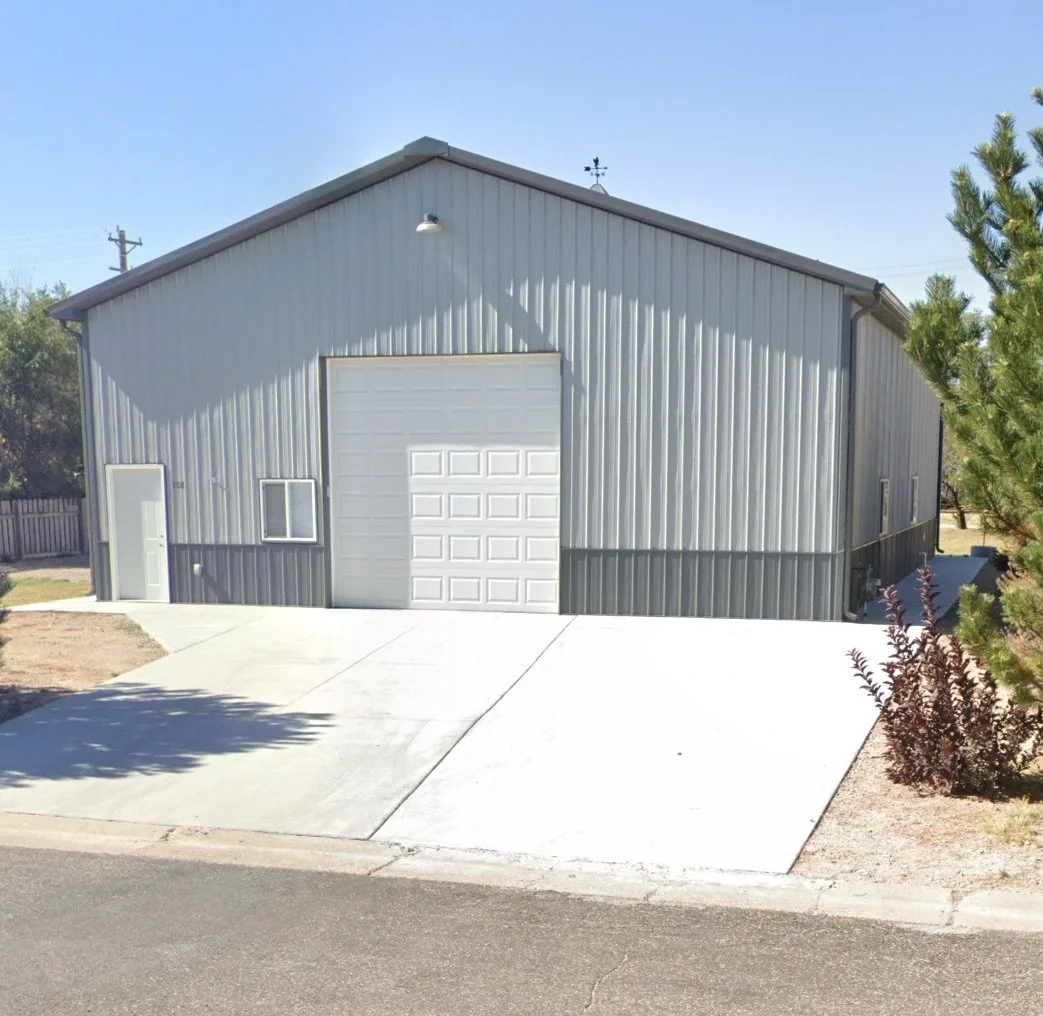 A large metal building used as a garage or storage shed, with a white roll-up door and a side entry door, situated on a concrete driveway and surrounded by some trees and bushes under a clear blue sky.