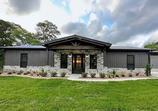 Front view of a modern single-story house with a combination of stone and dark metal siding, surrounded by a well-maintained lawn and trees, under a cloudy sky.