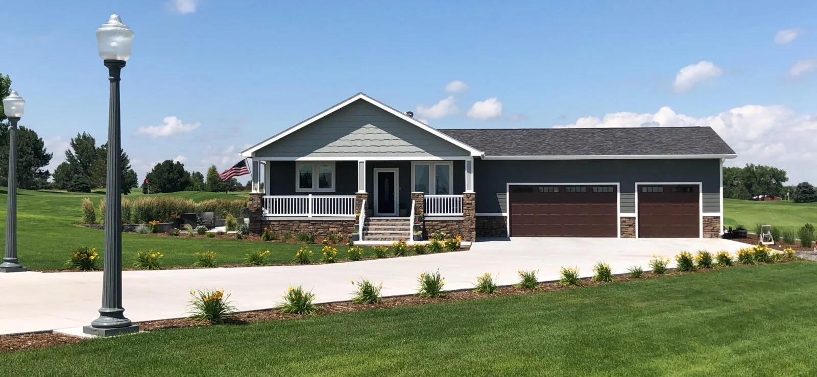 A house with gray exterior, front porch, two-car garage, surrounded by a well-maintained lawn and flower beds, under a blue sky with scattered clouds.