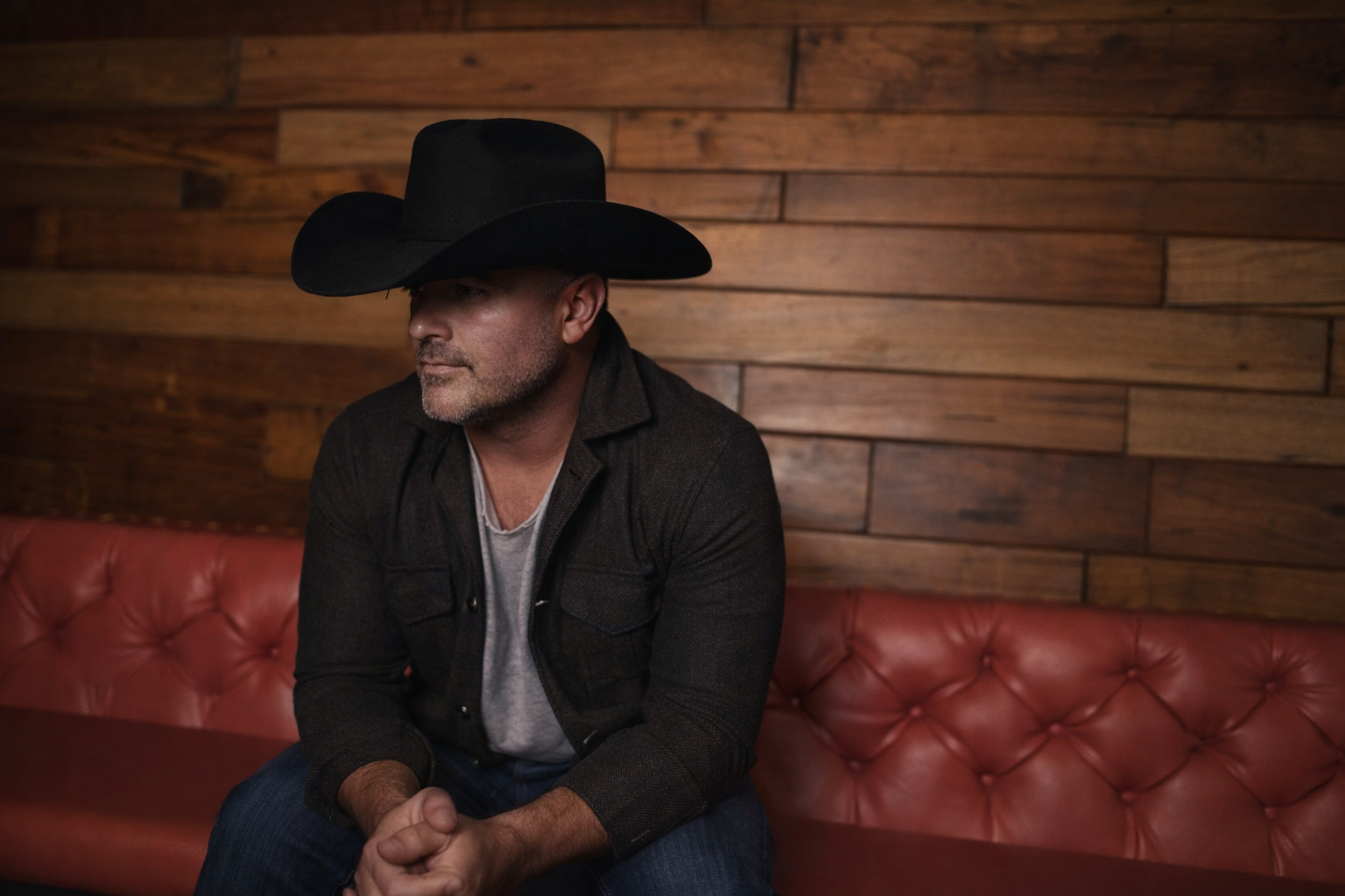 A man wearing a black cowboy hat, black jacket, and gray shirt sitting on a red leather tufted bench against a wooden wall.