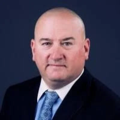 Professional headshot of a bald man in a suit and tie against a dark background.