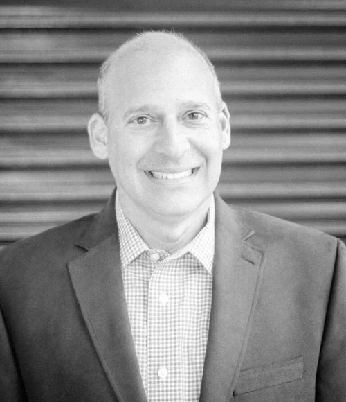 Black and white portrait of a smiling man wearing a business suit and checkered shirt, standing in front of horizontal wooden paneling.