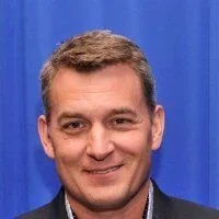 A man with short brown hair smiling in front of a blue curtain.