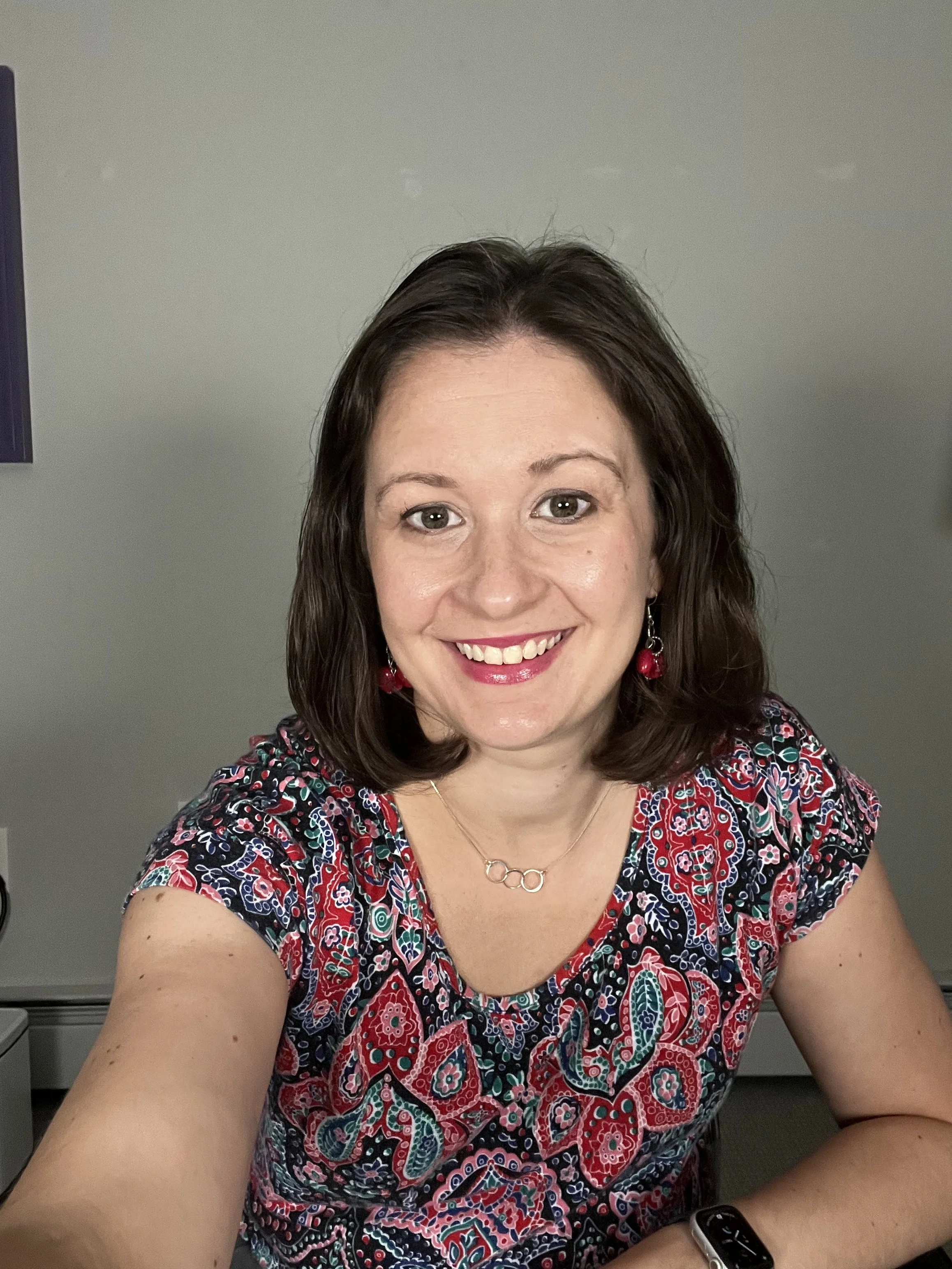 A woman with shoulder-length brown hair smiling at the camera, wearing a colorful patterned top, red earrings, a silver necklace with three rings, and a fitness tracker on her left wrist, sitting at a desk in an office with a gray wall background.