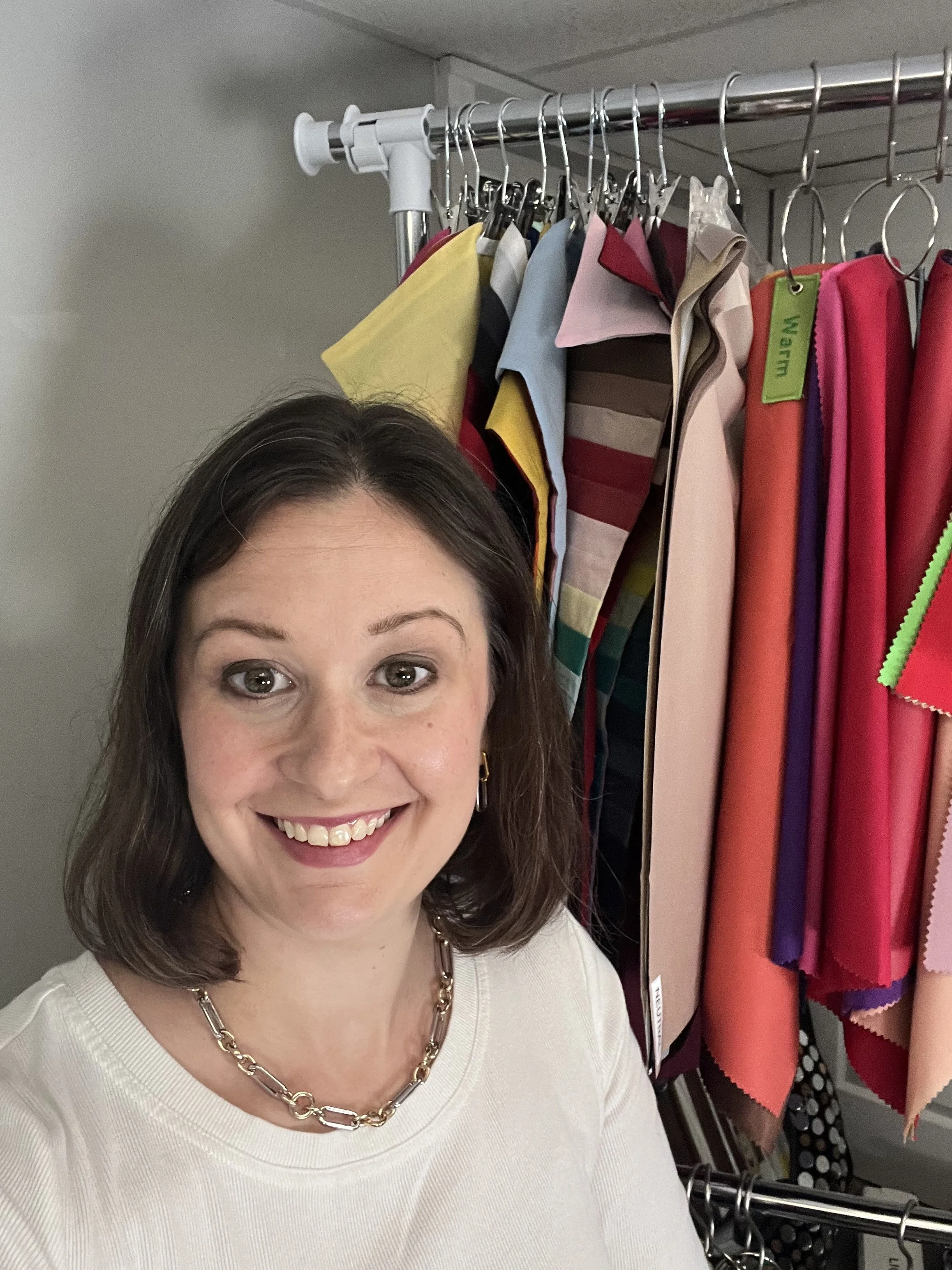 A woman with dark brown hair and light skin smiling in front of a clothing rack with fabrics and textiles.