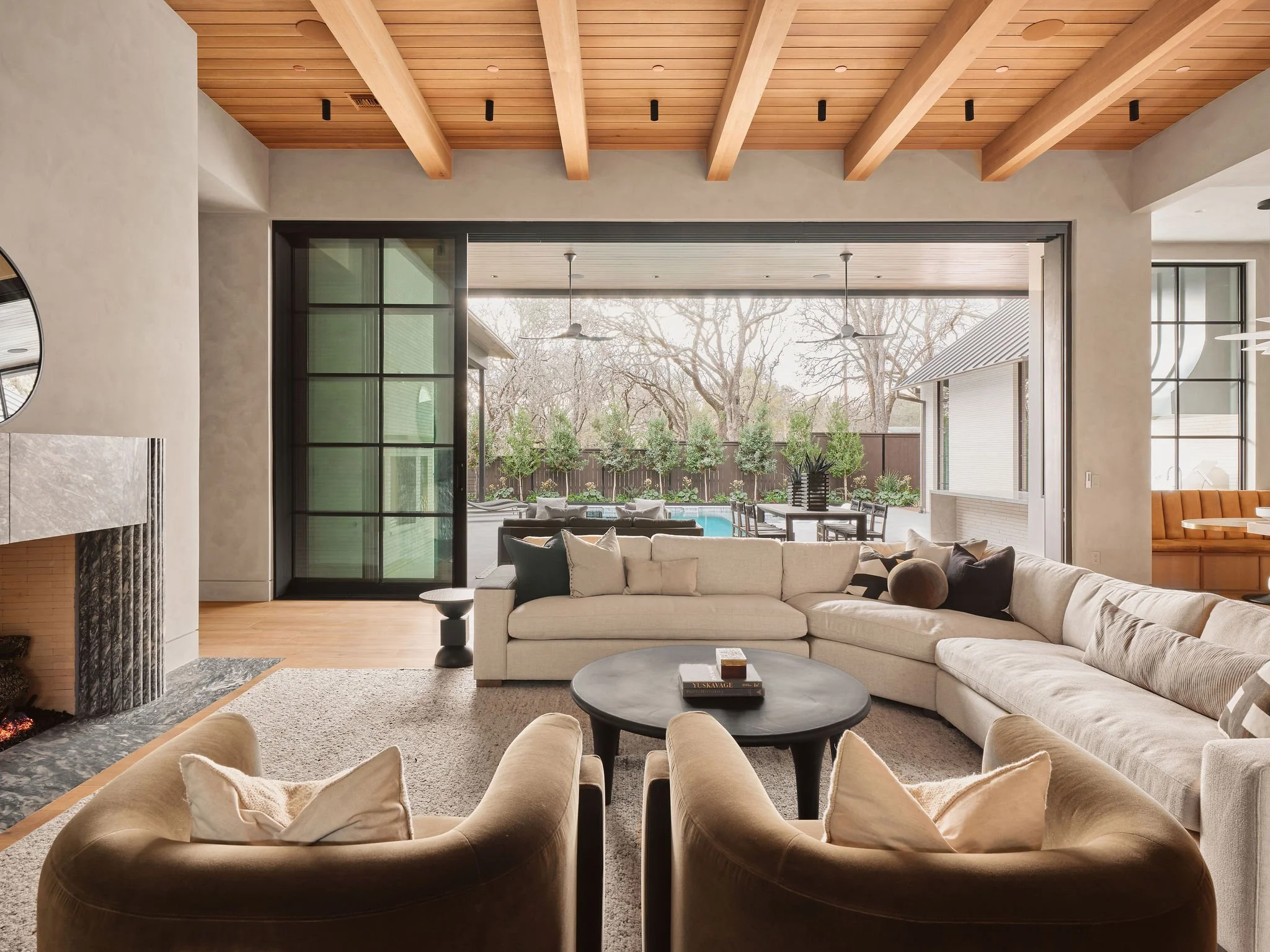 Modern living room with large beige sectional sofa, black round coffee table, and beige armchairs in front of a fireplace, with large windows and a patio with trees visible outside.
