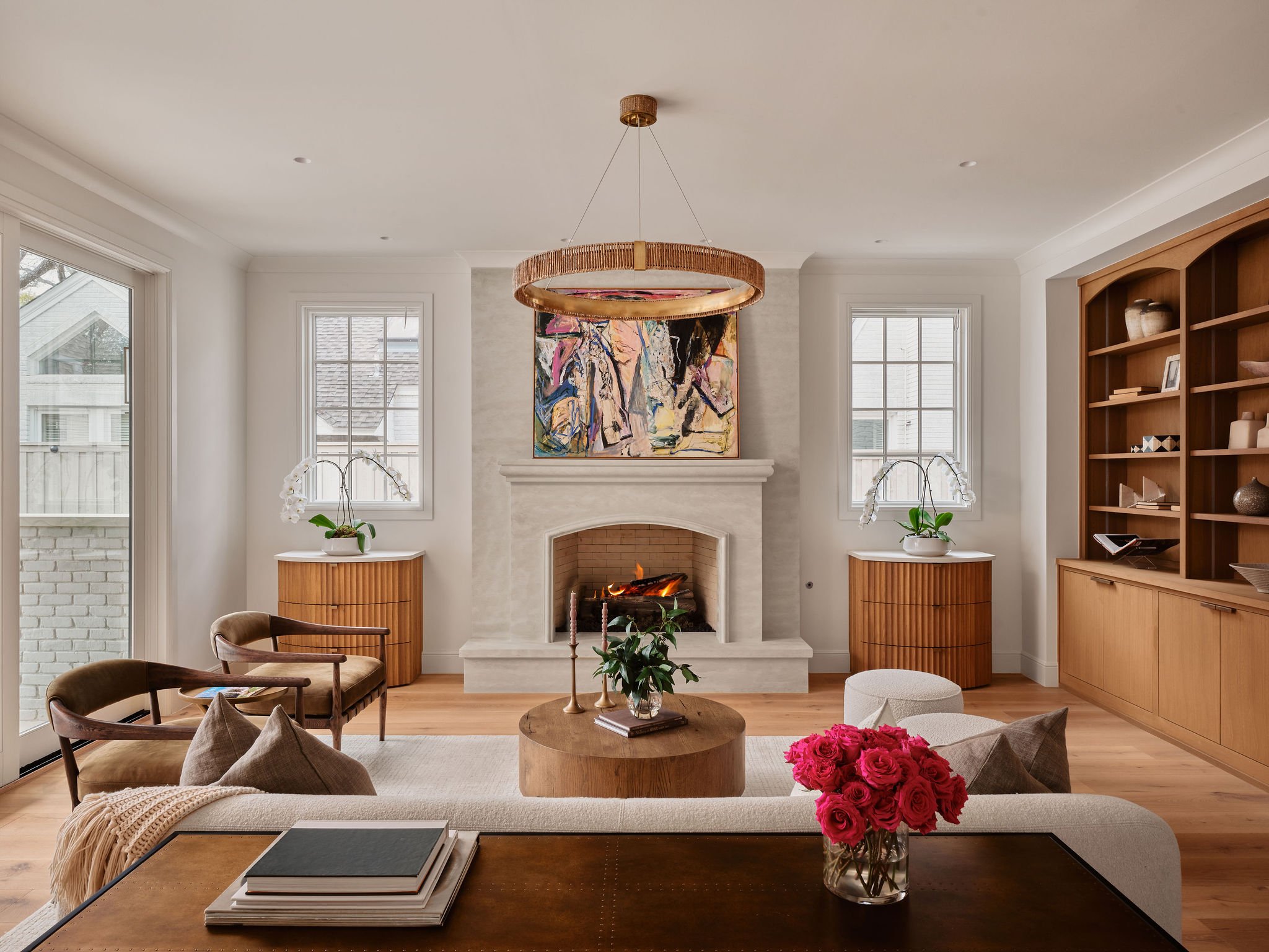 A bright living room with a white fireplace, abstract painting above it, large windows on either side, and wooden furniture including a bookshelf and side tables. There are chairs, a vase with pink flowers, and a round coffee table with books and candles.