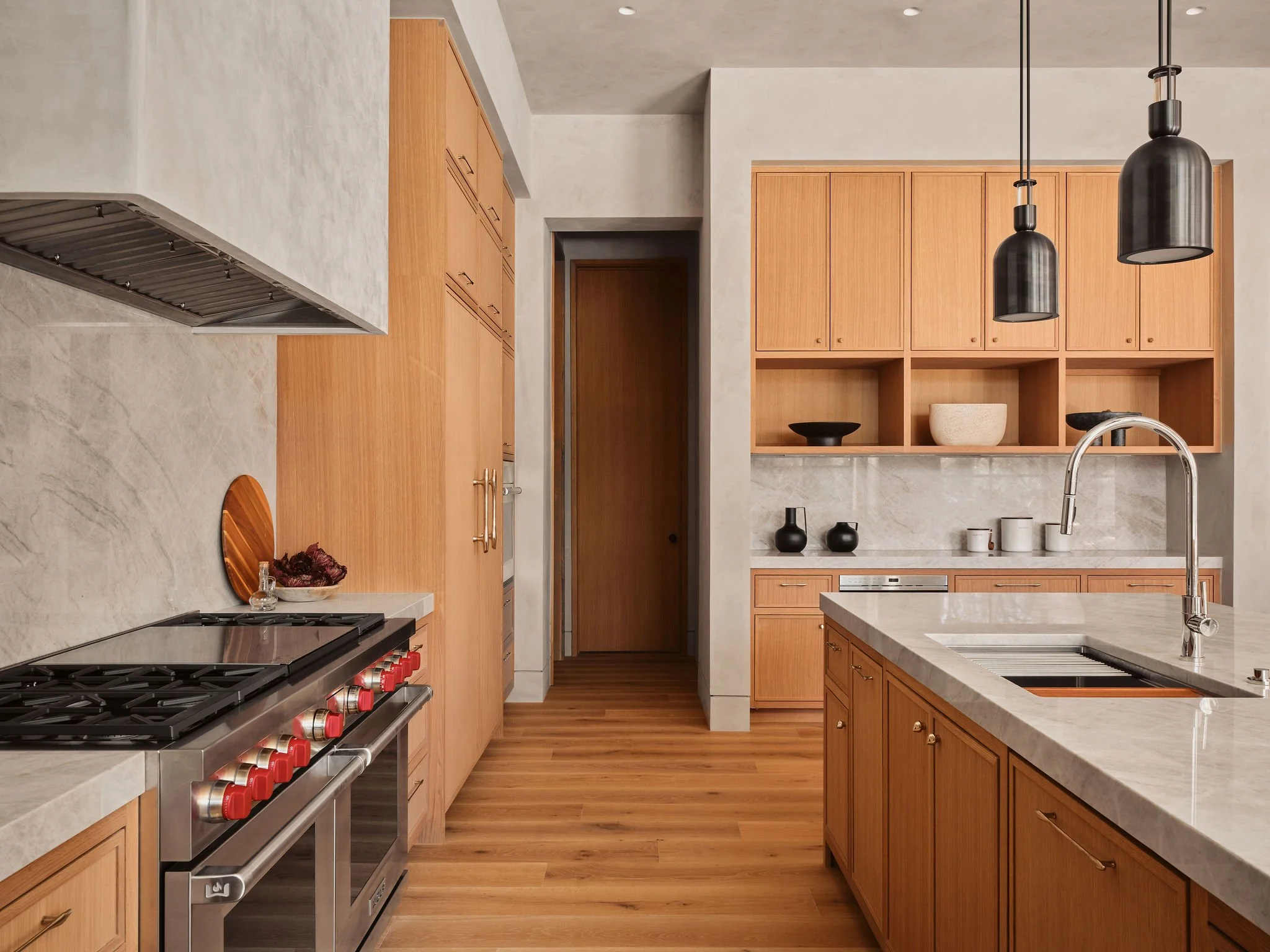Modern kitchen with wooden cabinets, marble countertops, a stainless steel stove with red knobs, and two black pendant lights hanging from the ceiling.