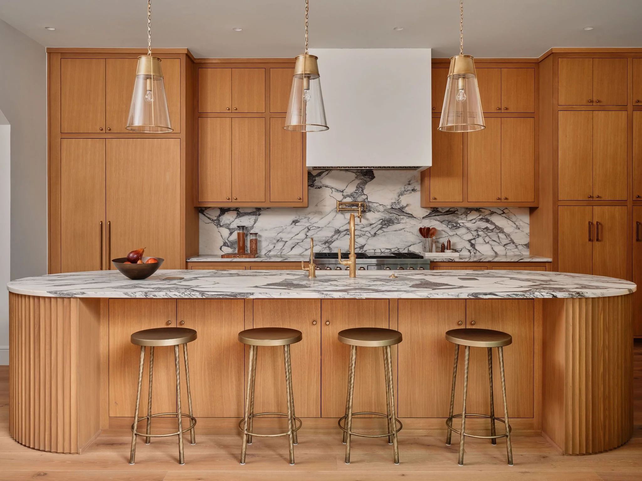Kitchen with wooden cabinetry, marble backsplash, three pendant lights, and a marble kitchen island with four bar stools.
