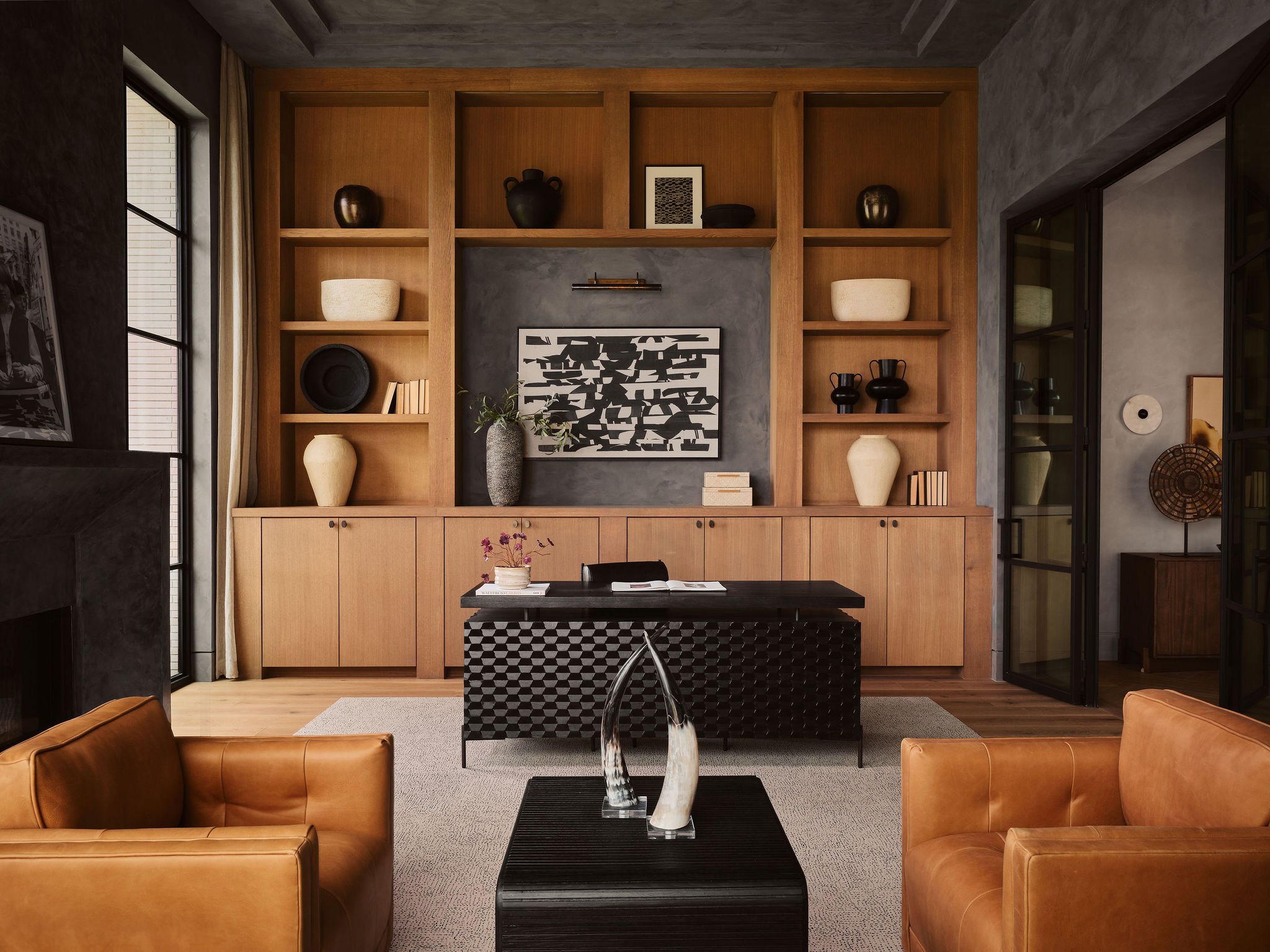 Modern living room with a built-in wooden bookshelf, abstract artwork, decorative vases, and a wooden cabinet. Brown leather armchairs and a black coffee table with sculptures are visible. Large windows with curtains are on either side.