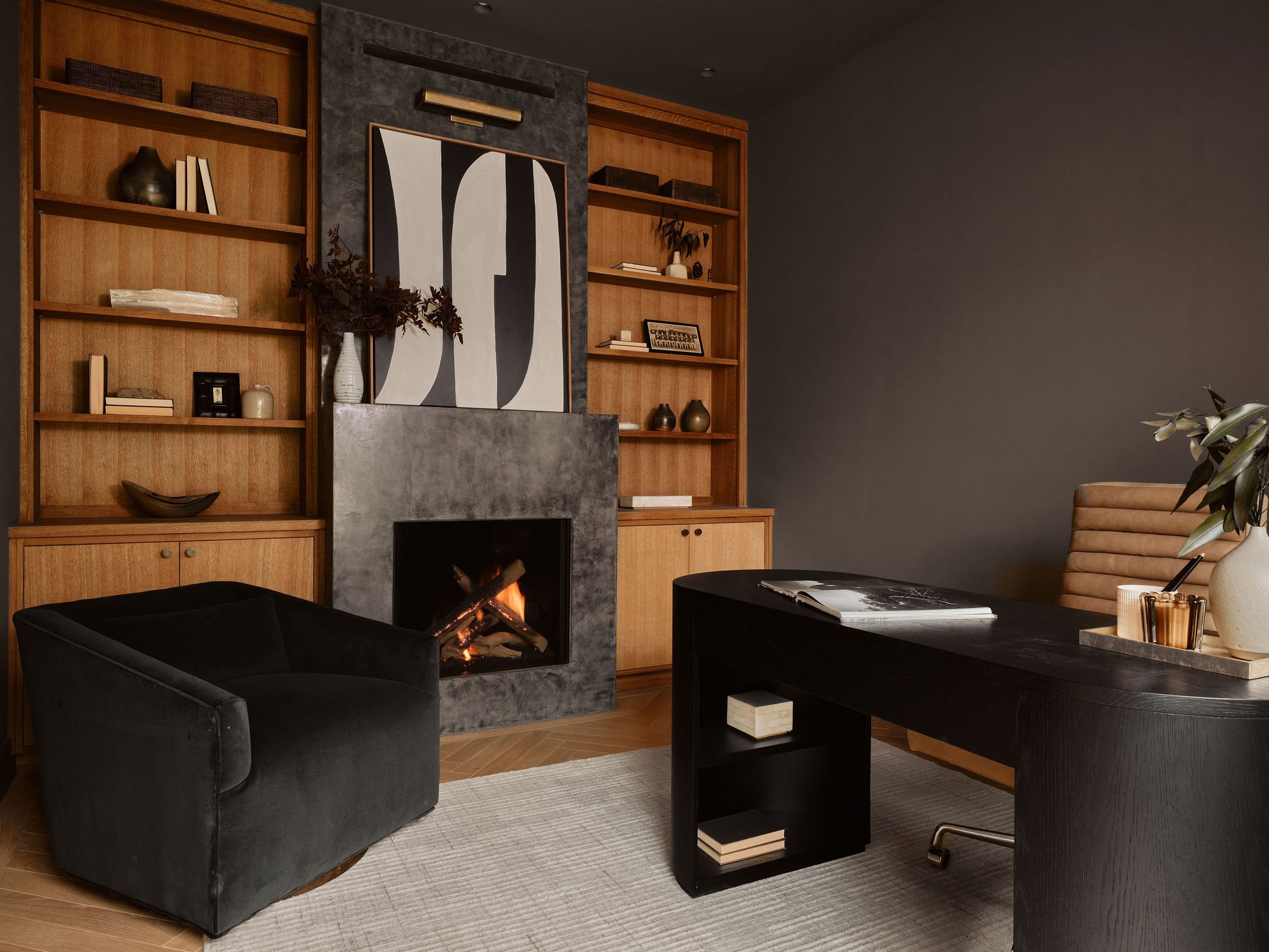 Modern living room with a built-in wooden bookcase, a fireplace with abstract black-and-white artwork, black armchair, dark wooden coffee table with magazines, and a beige armchair with a plant nearby.