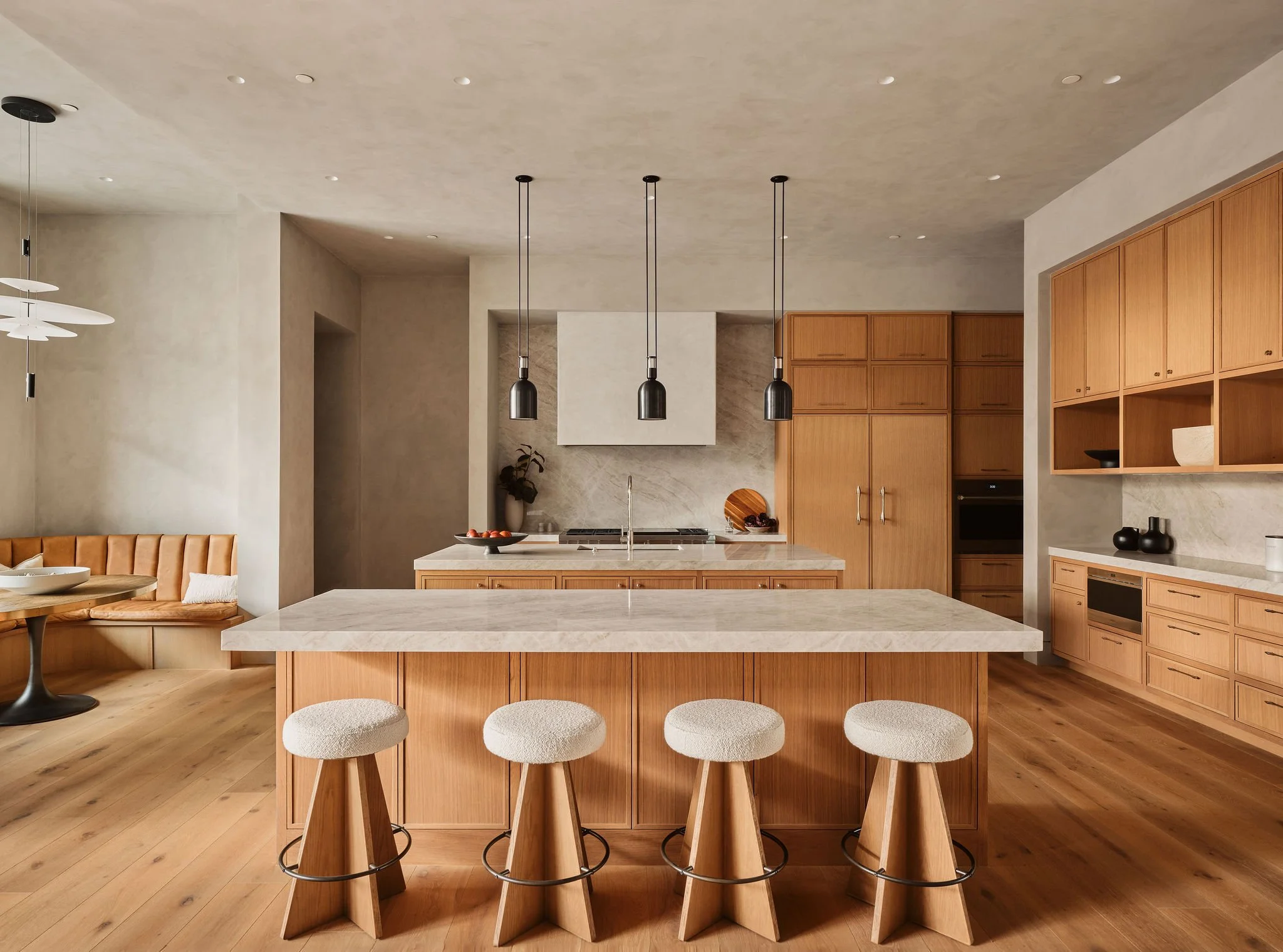A modern kitchen with a large island, three black pendant lights hanging above, and natural wood cabinetry. A row of padded stools lines the front of the island. The kitchen features neutral tones and minimal decor.