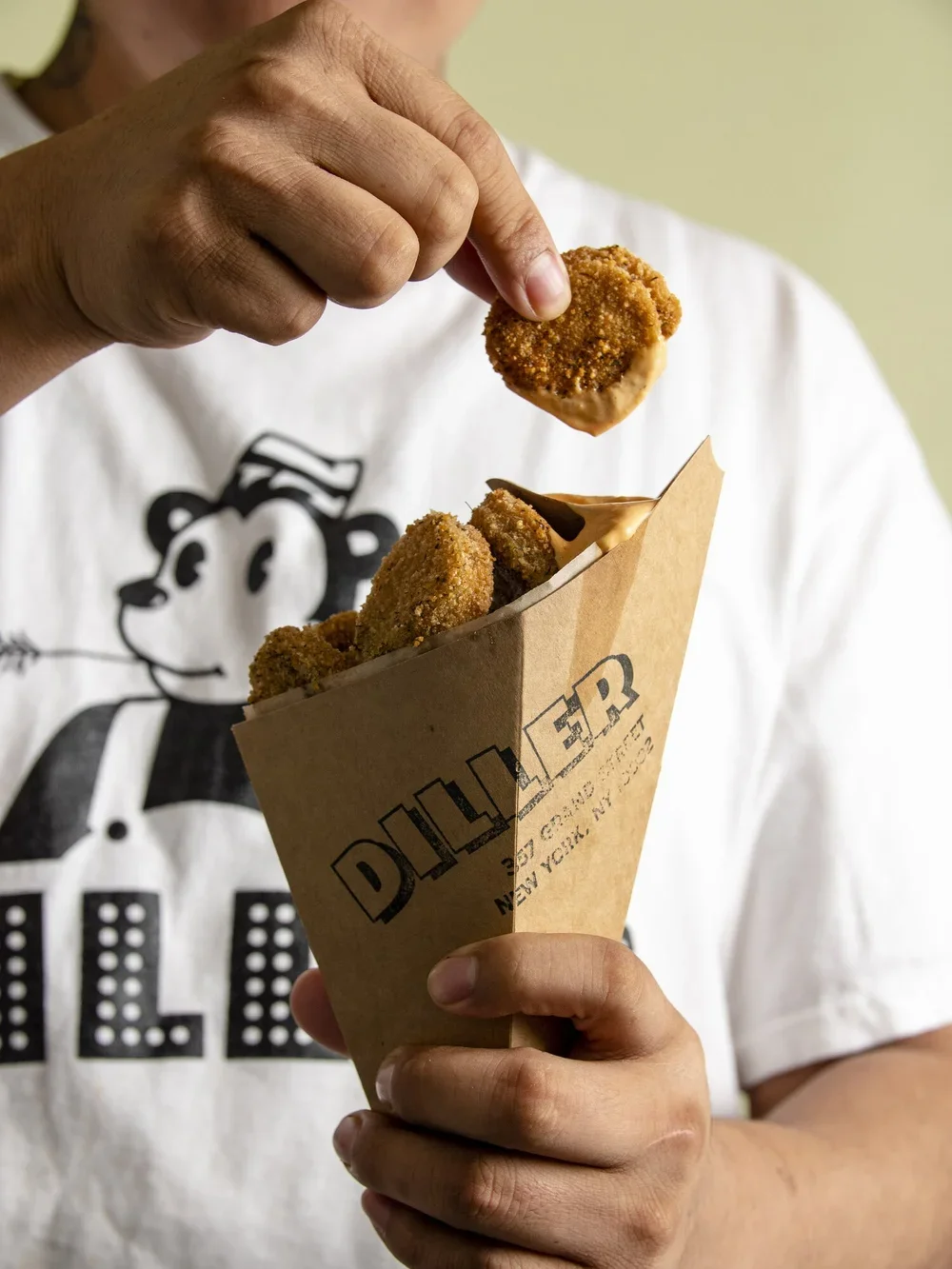 Person holding a paper cone filled with breaded and fried chicken nuggets, with someone dipping one nugget into a sauce.