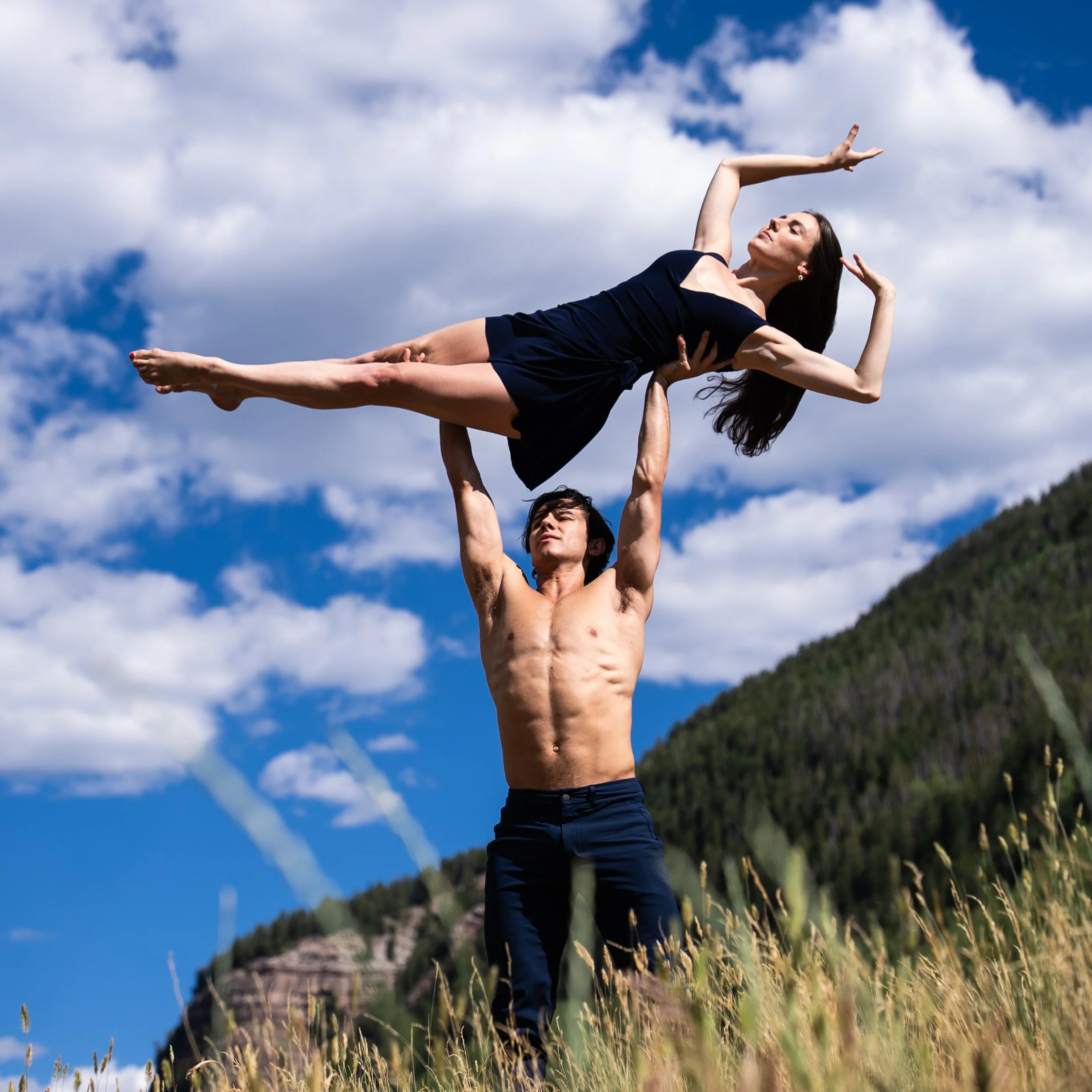 Tiler Peck and Roman Mejia dance outdoors against a backdrop of blue sky with scattered clouds and a green hill at the Vail Dance Festival in Colorado.