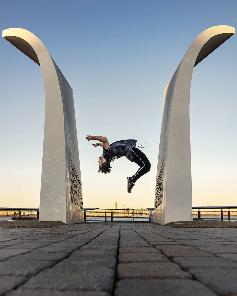 A person performing a backflip between two large, white, curved monument structures on a paved walkway with a city skyline in the distance during sunset.