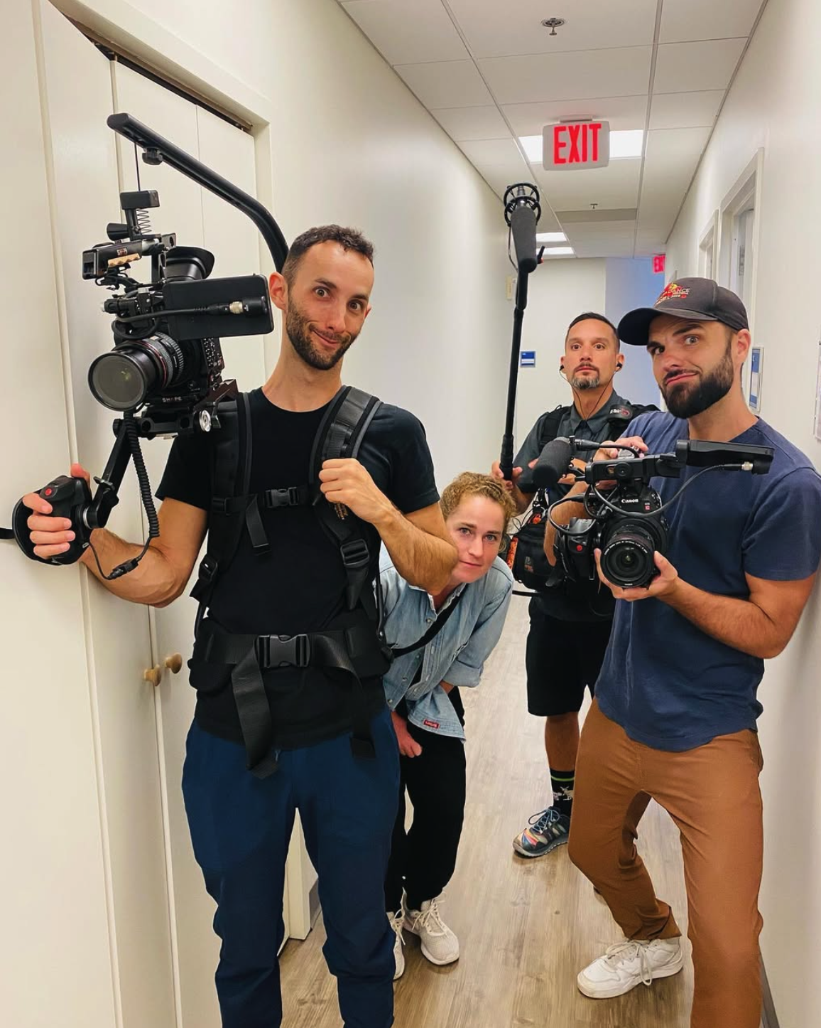 Nate Reininga and Cody Buesing holding professional video cameras. They appear to be filming or preparing to film, with one person holding a boom microphone. The hallway has white walls, a closed door, and an illuminated exit sign on the ceiling.