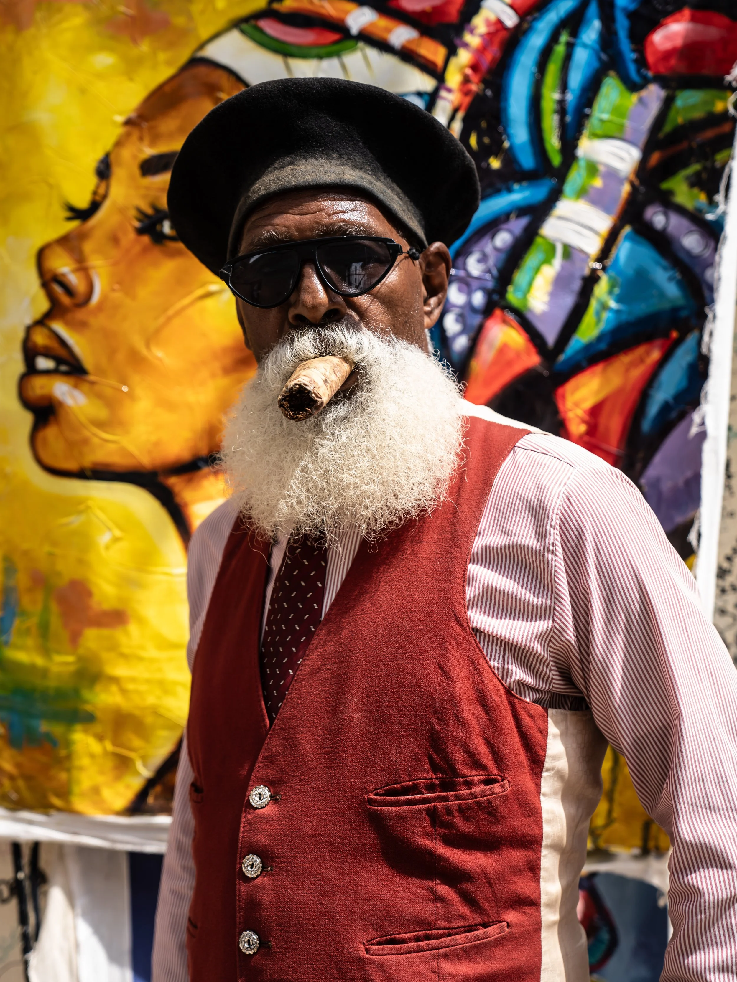 A man with a white beard wearing sunglasses, a black hat, and a red vest over a striped shirt, also smoking a cigar, standing in front of colorful street art.