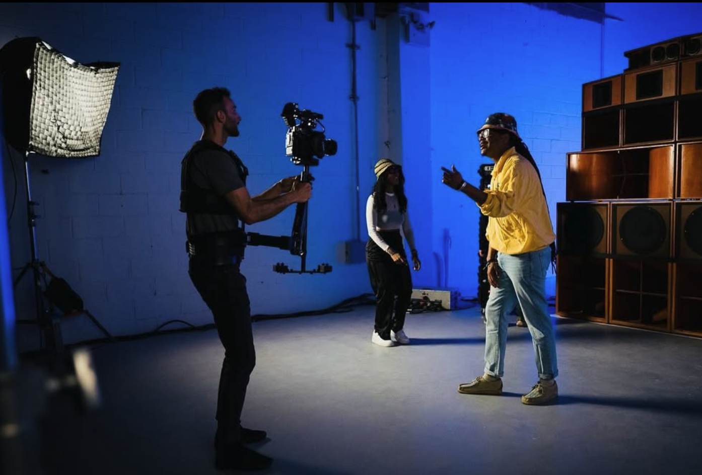 People filming a scene in a studio with blue lighting, large speakers, and professional filming equipment.
