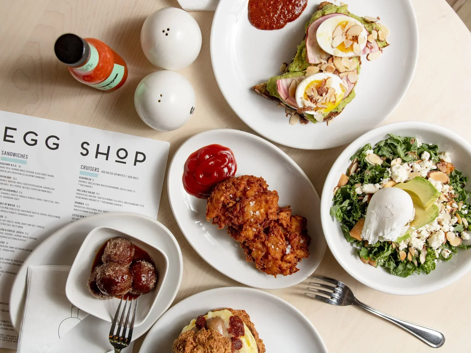A table with various dishes including a salad with avocado, hard-boiled eggs, and nuts, a piece of fried chicken with ketchup, a plate with meatballs and sauce, and an open-faced sandwich with avocado, egg, and sliced almonds, along with salt and pepper shakers, hot sauce, and a menu from the Egg Shop.