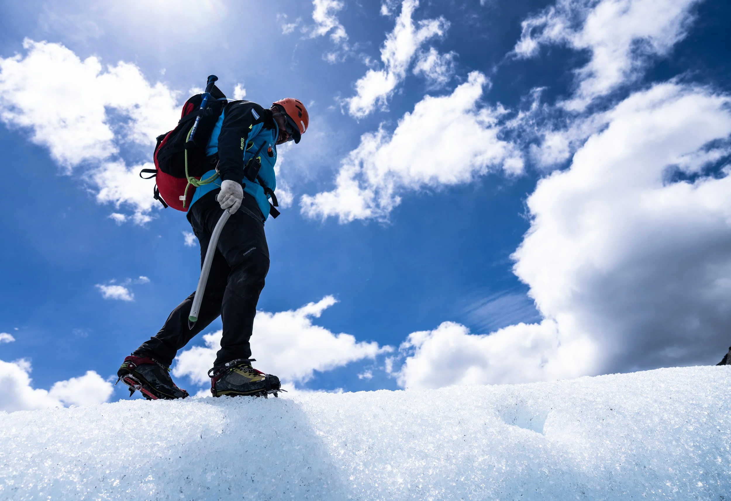 A person wearing a helmet and winter gear, standing on snow with climbing or hiking poles, under a partly cloudy sky.
