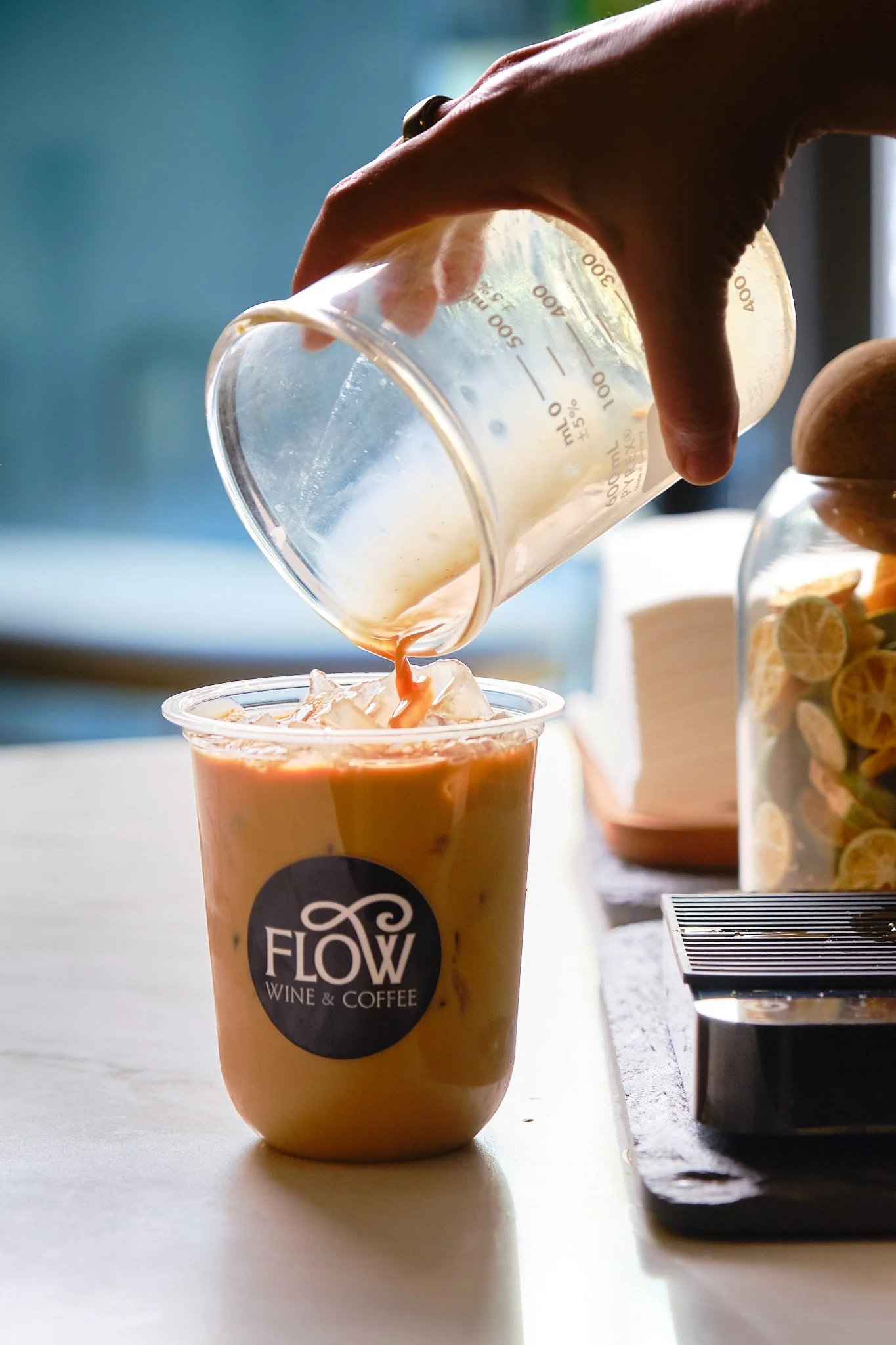 Person pouring a creamy beverage from a measuring cup into a plastic cup branded 'Flow Wine & Coffee'; in the background, a jar of lemons and a container of sliced lemons are visible.