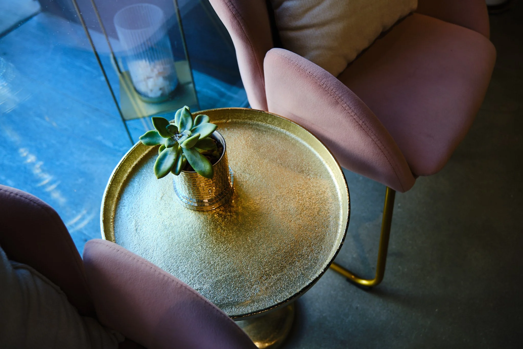 A small green succulent plant in a small gold-colored pot on a round, shiny, gold-colored table. The table and plant are next to a light pink upholstered chair with a curved armrest. The background shows a glass surface with a reflection and a blurre